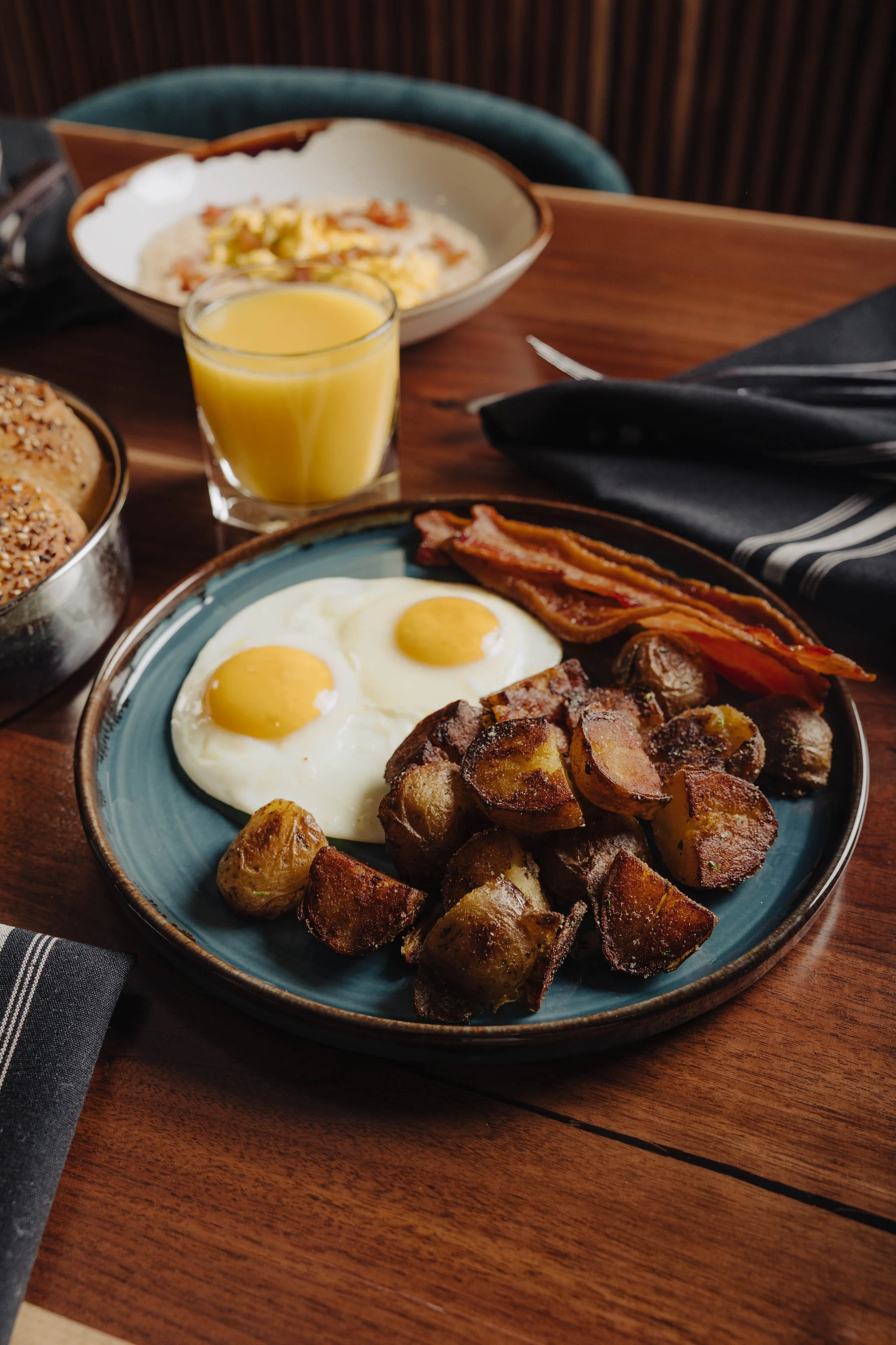 A breakfast plate with two sunny side up eggs, bacon, roasted potatoes, and sausage links. There is a glass of orange juice and other breakfast items in the background.