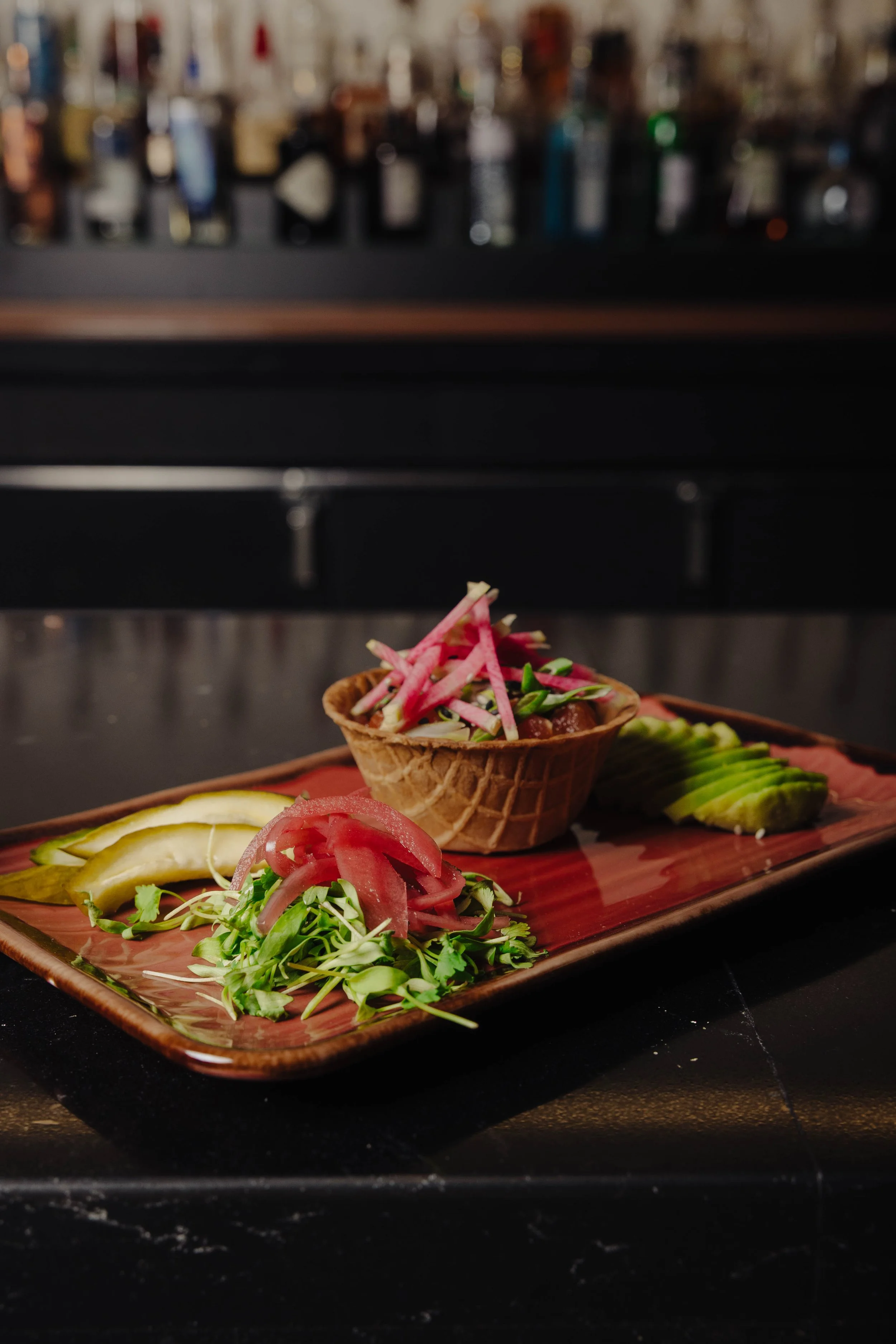 A rectangular brown ceramic plate with sliced avocado, shredded radish and herbs, and a small waffle cup filled with a salad topped with radish strips, set against a blurred bar background.
