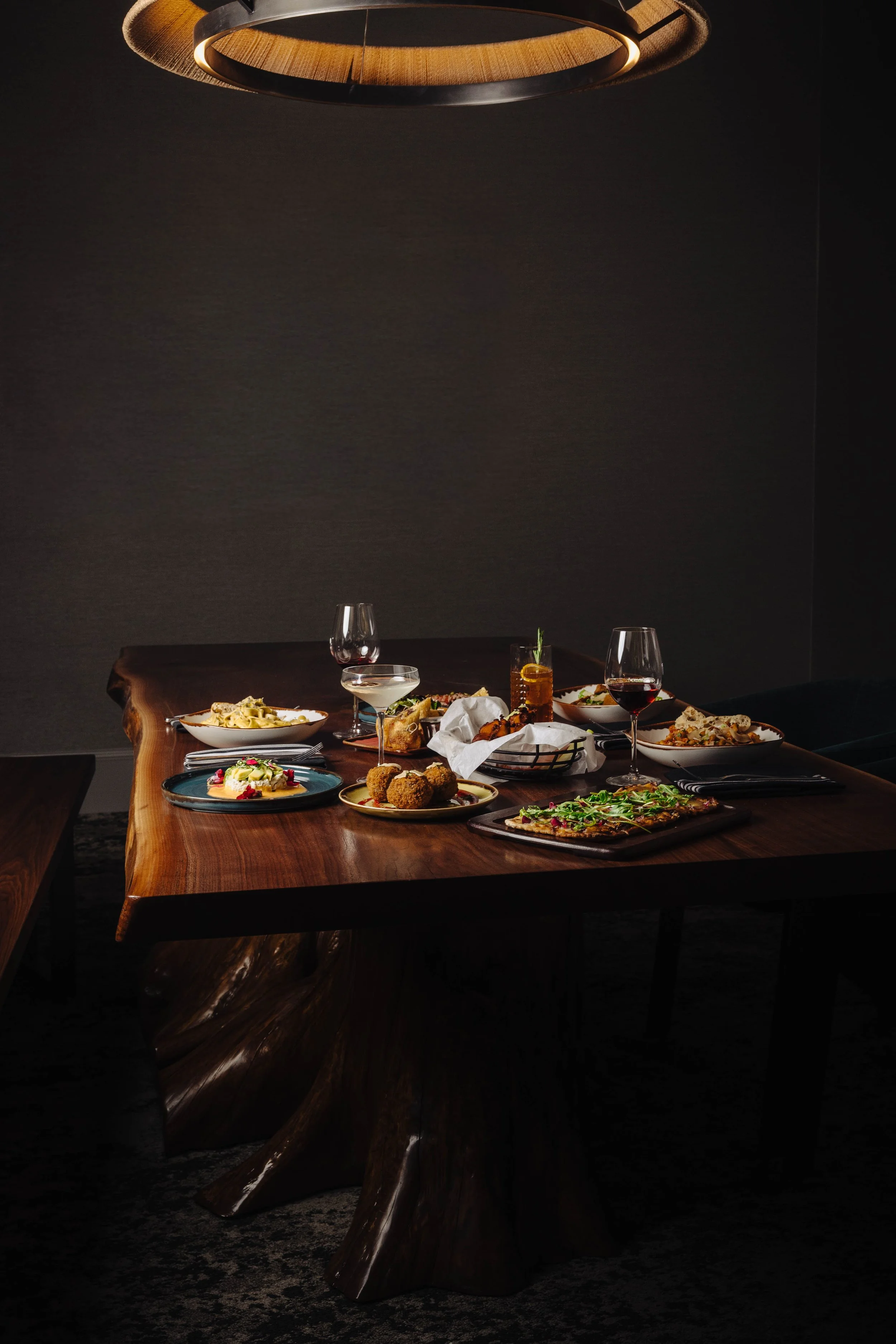 A wooden dining table set with various dishes and drinks, illuminated by a modern overhead light, with a dark background.