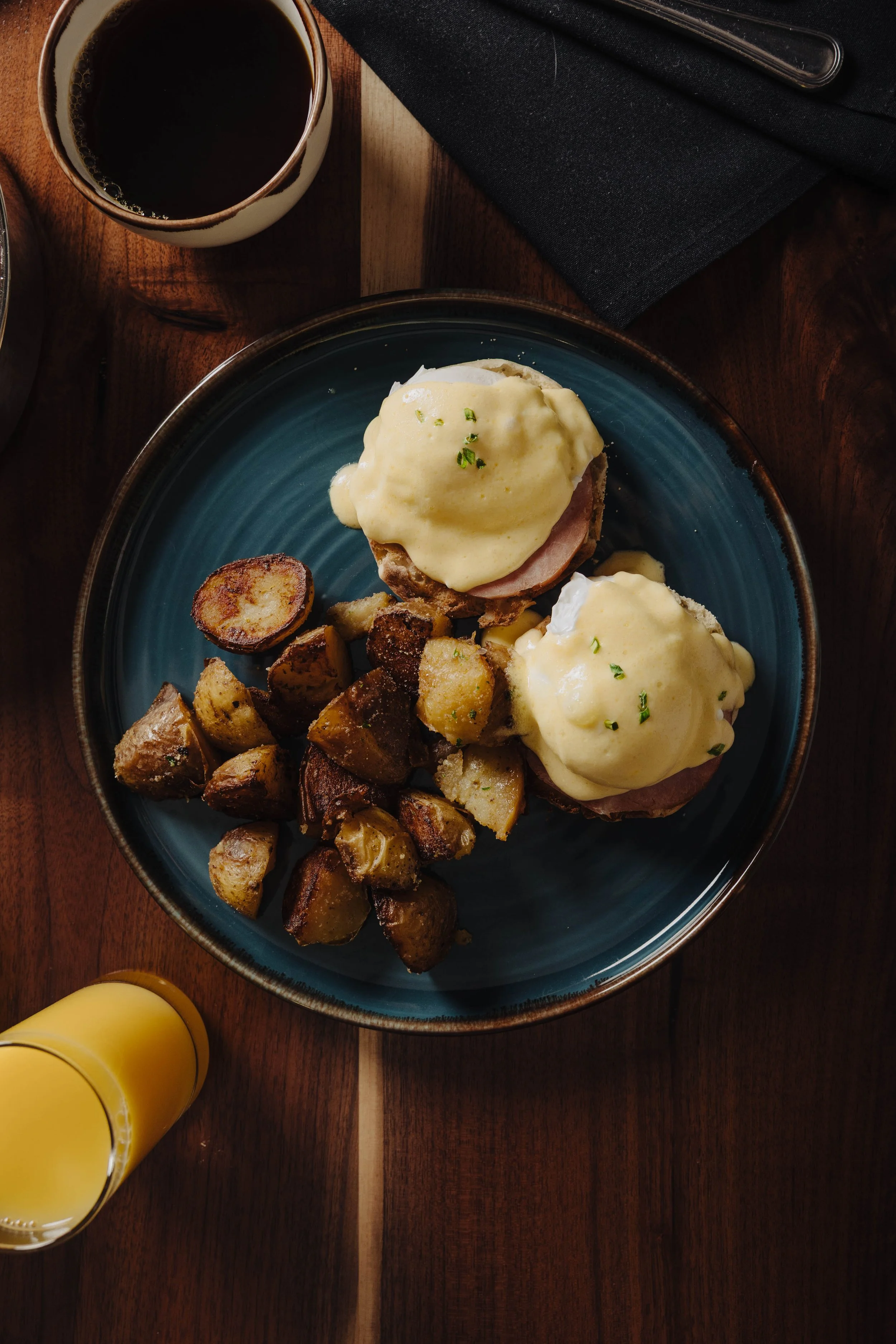 Plate with two eggs Benedict with hollandaise sauce, roasted potatoes, a cup of coffee, and a glass of orange juice on a wooden table.