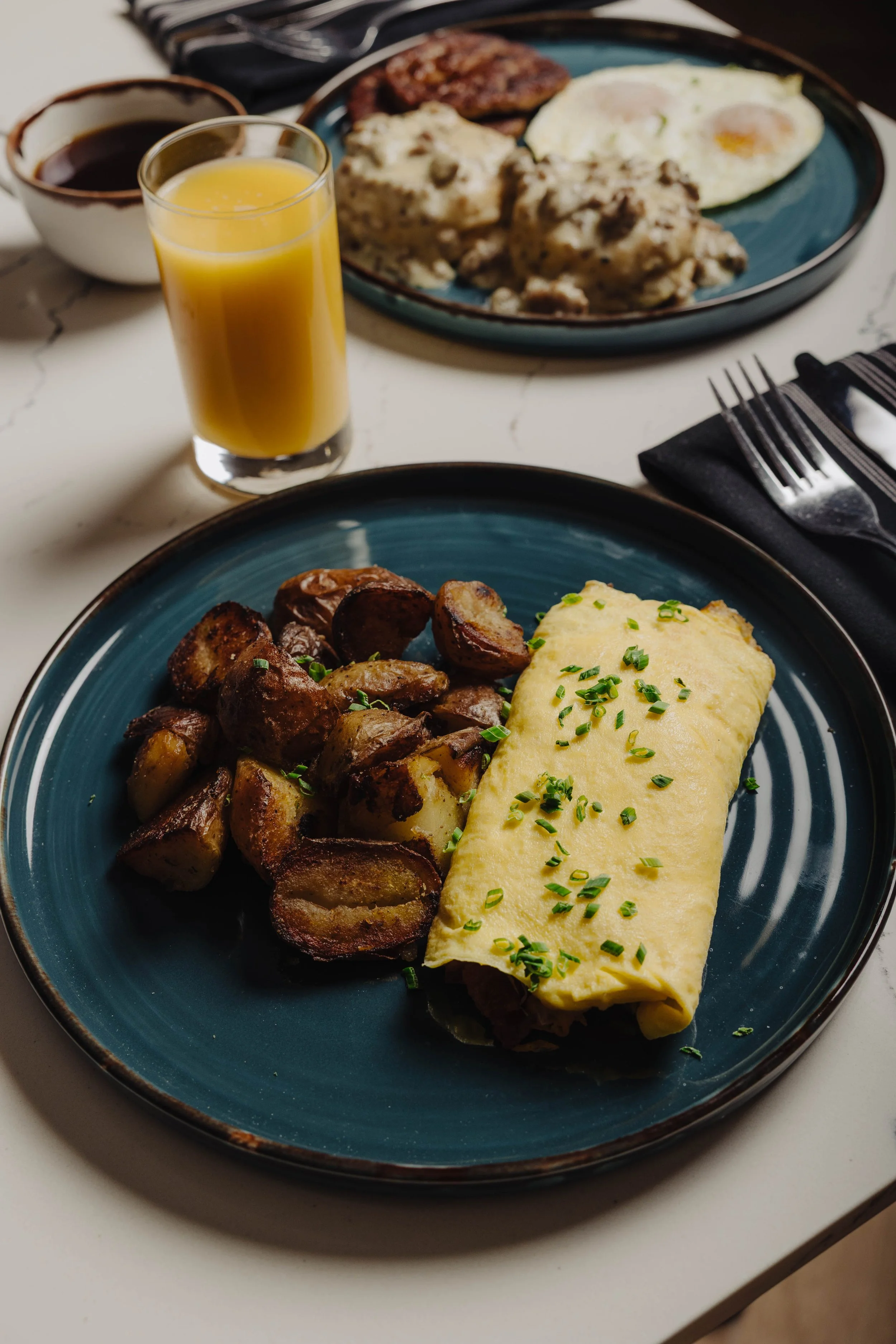 A breakfast plate with an omelet garnished with chopped chives, roasted potatoes, a glass of orange juice, and a side of sausage and eggs in the background.