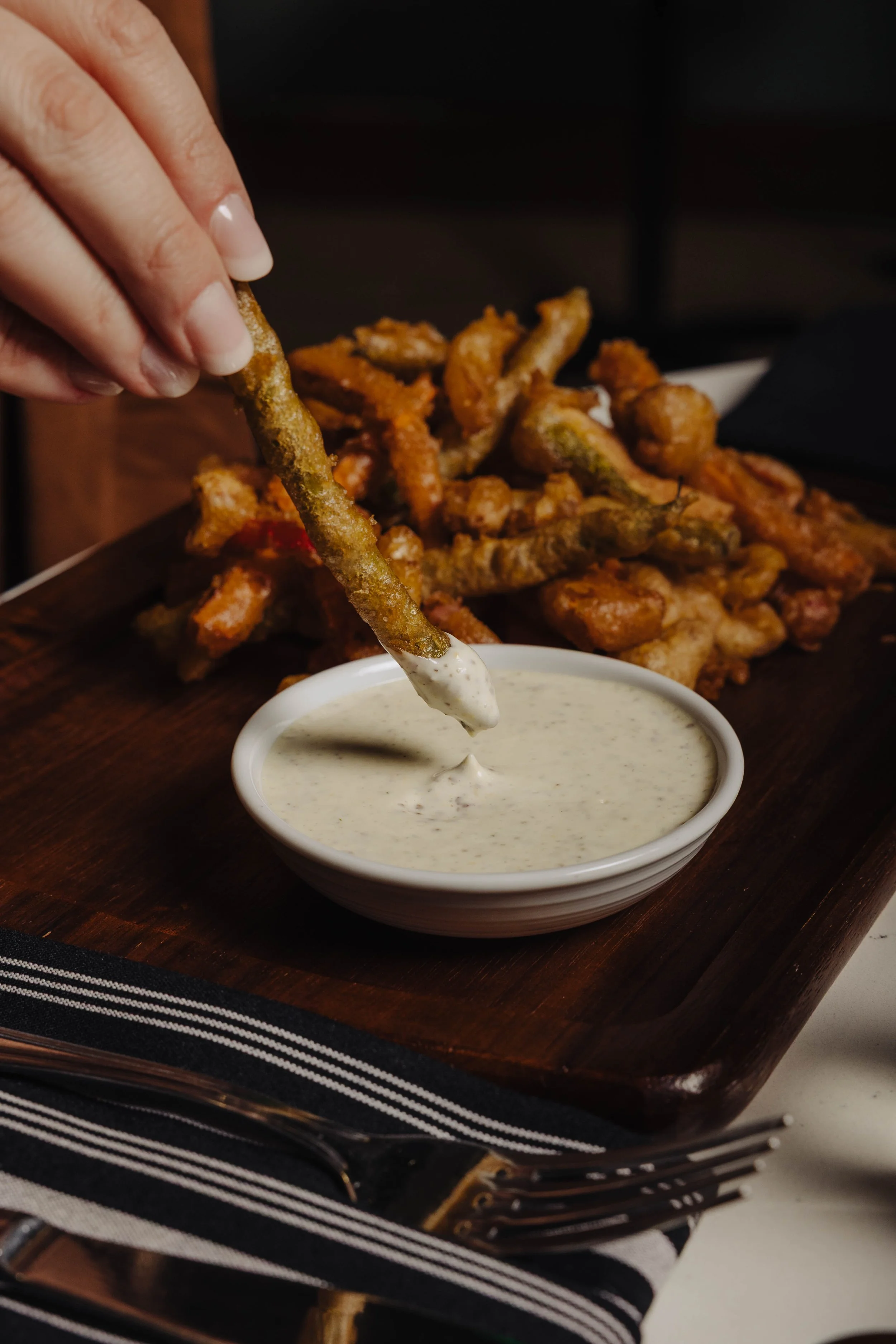 Fried green beans being dipped into a bowl of creamy dipping sauce, served on a wooden tray.