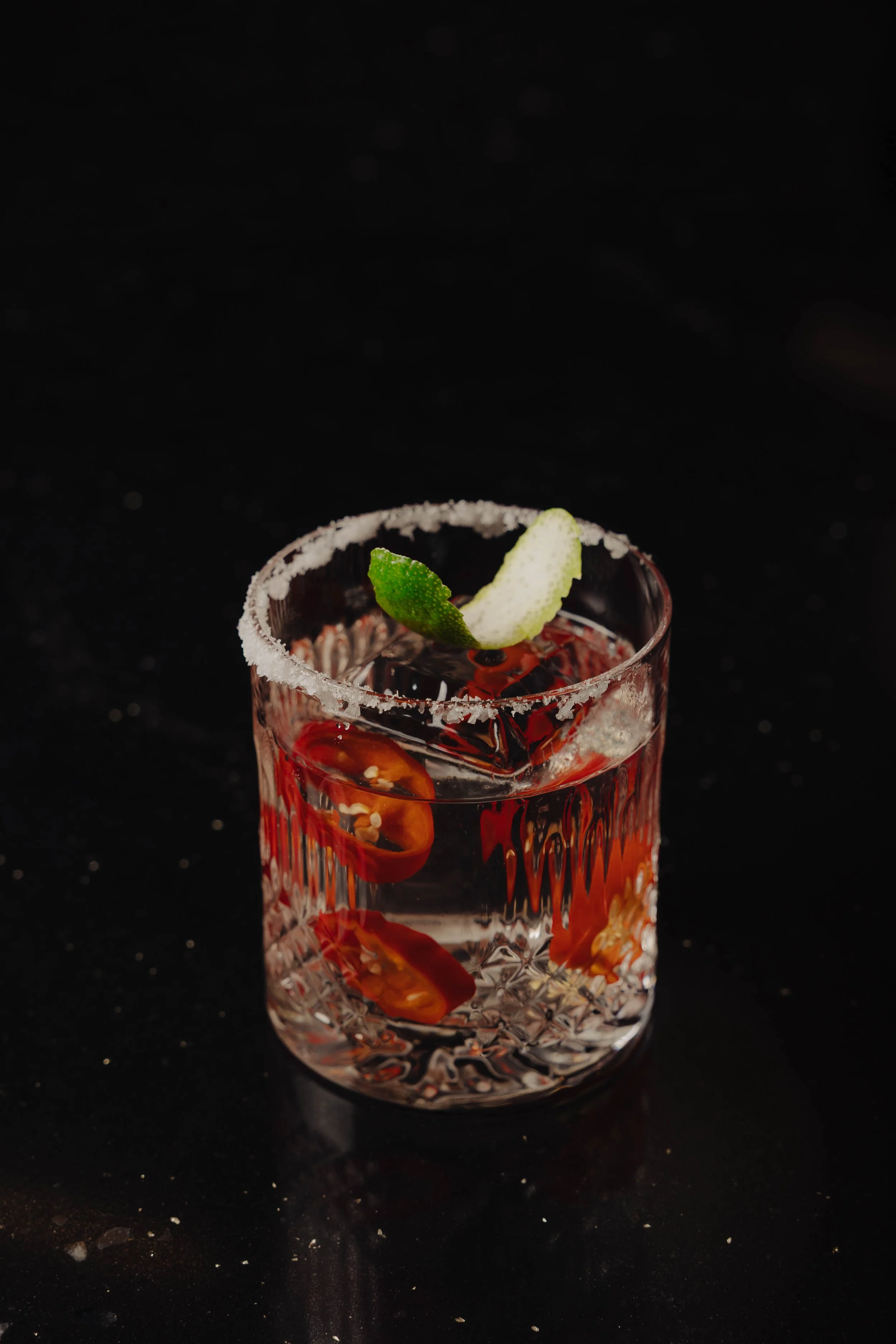 A glass filled with a clear beverage, garnished with a lime wedge and sliced red chili peppers, on a black surface.