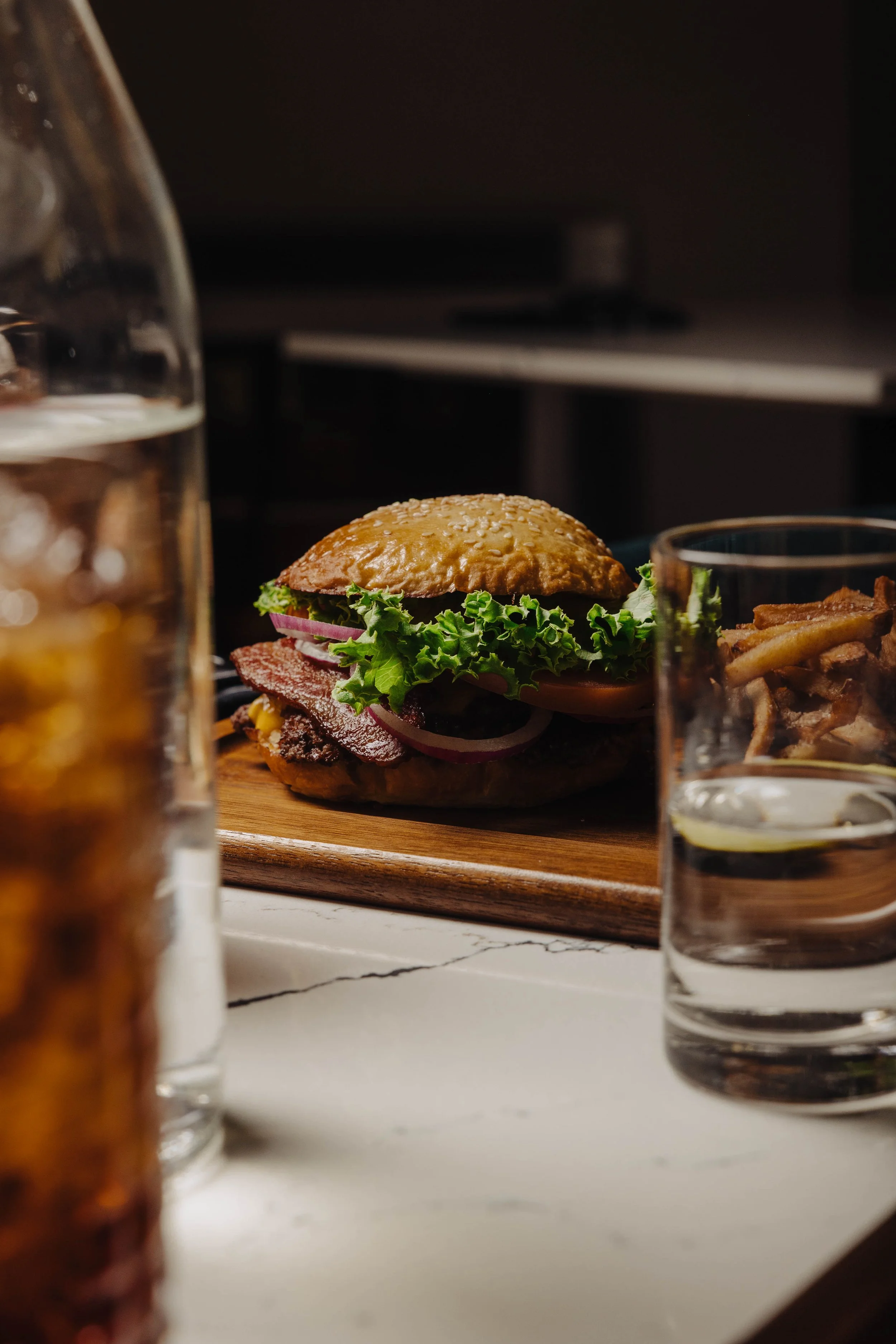 A burger with lettuce, tomato, onions, bacon, and a sesame seed bun on a wooden board, with glasses of water and a drink with ice in the foreground.