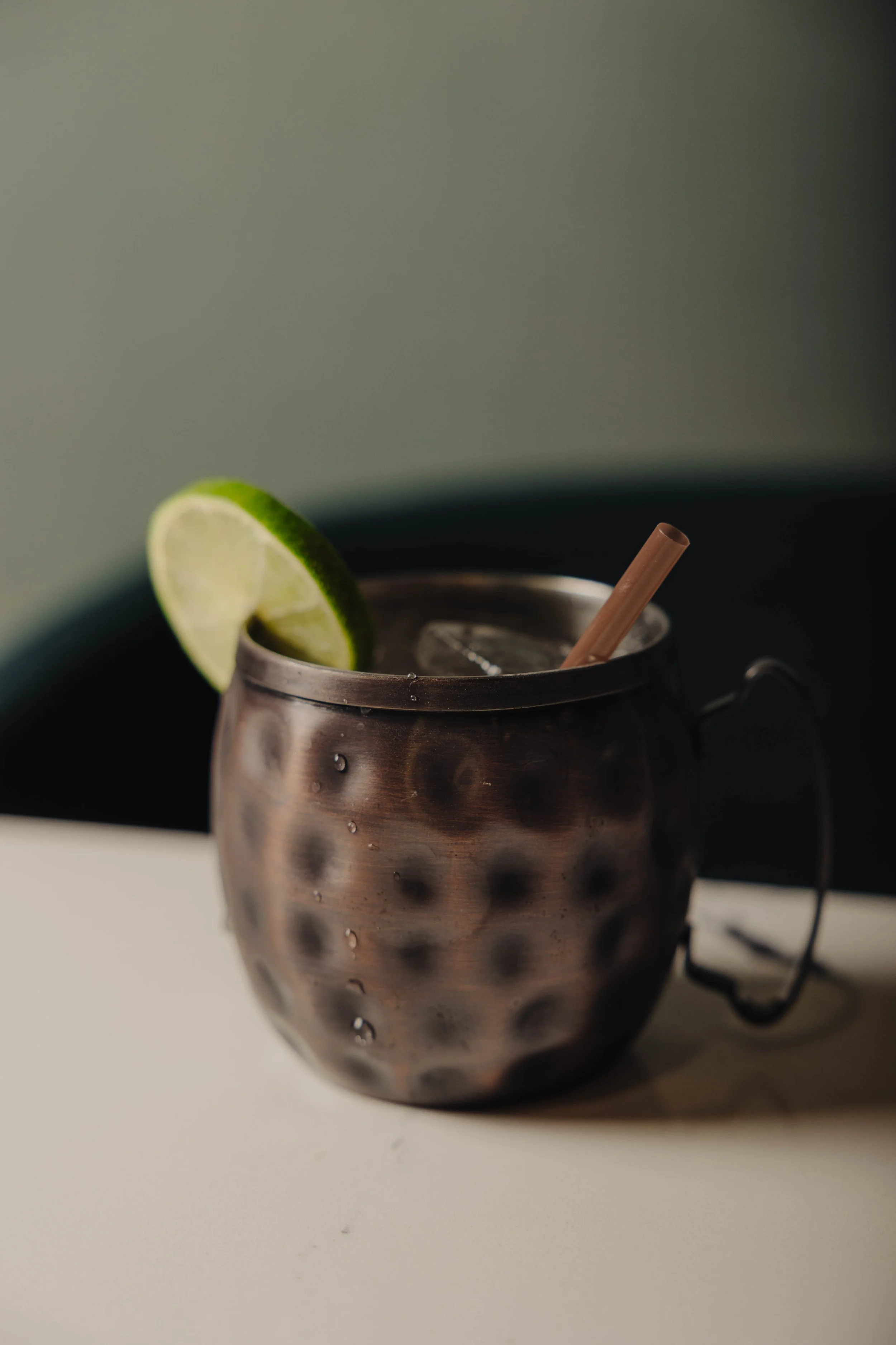 Metal cup with ice, a lime wedge, a brown straw, and condensation on the outside, sitting on a white surface against a dark background.