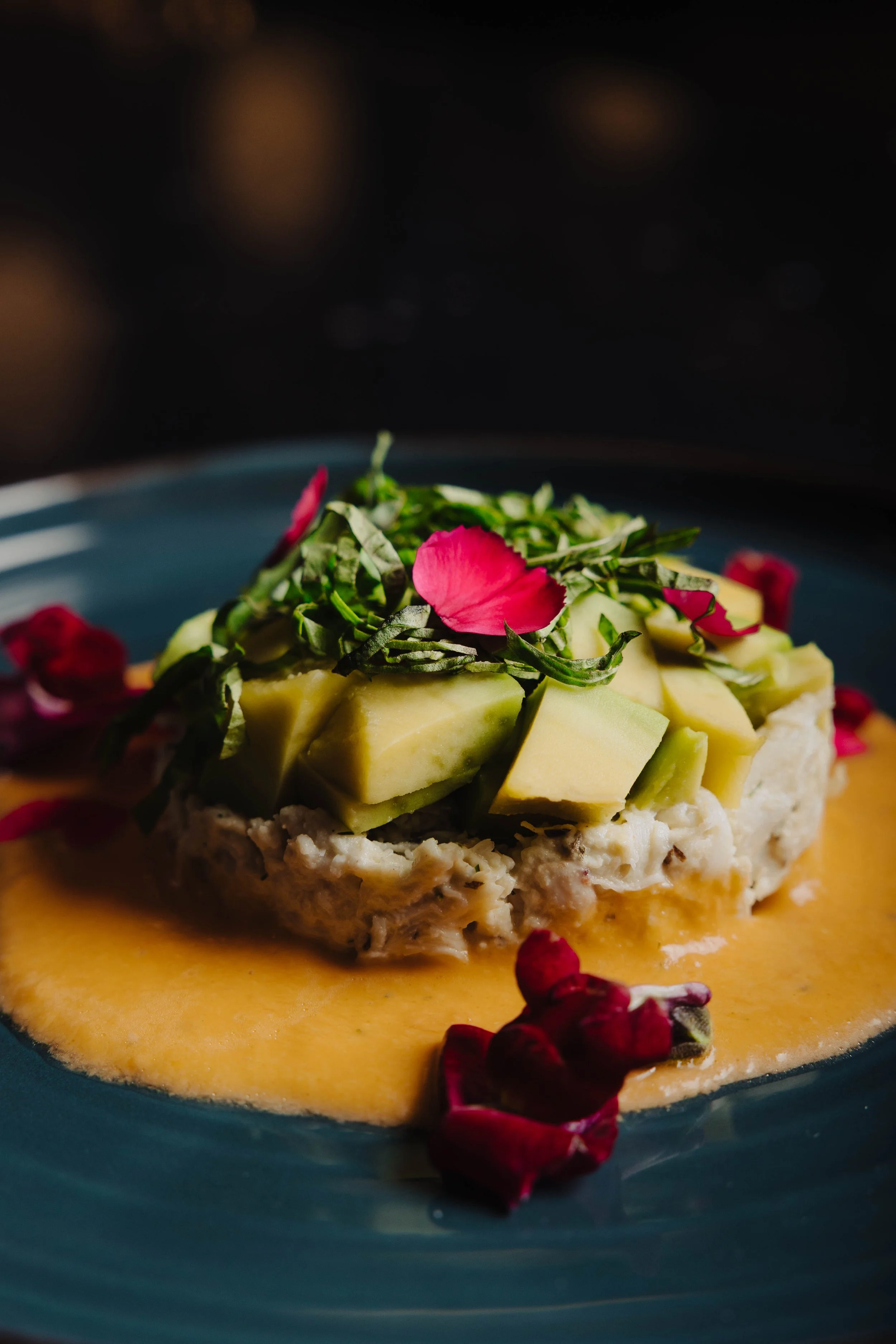 A dish with diced avocado, a sauce, and garnished with fresh herbs and pink edible flower petals.