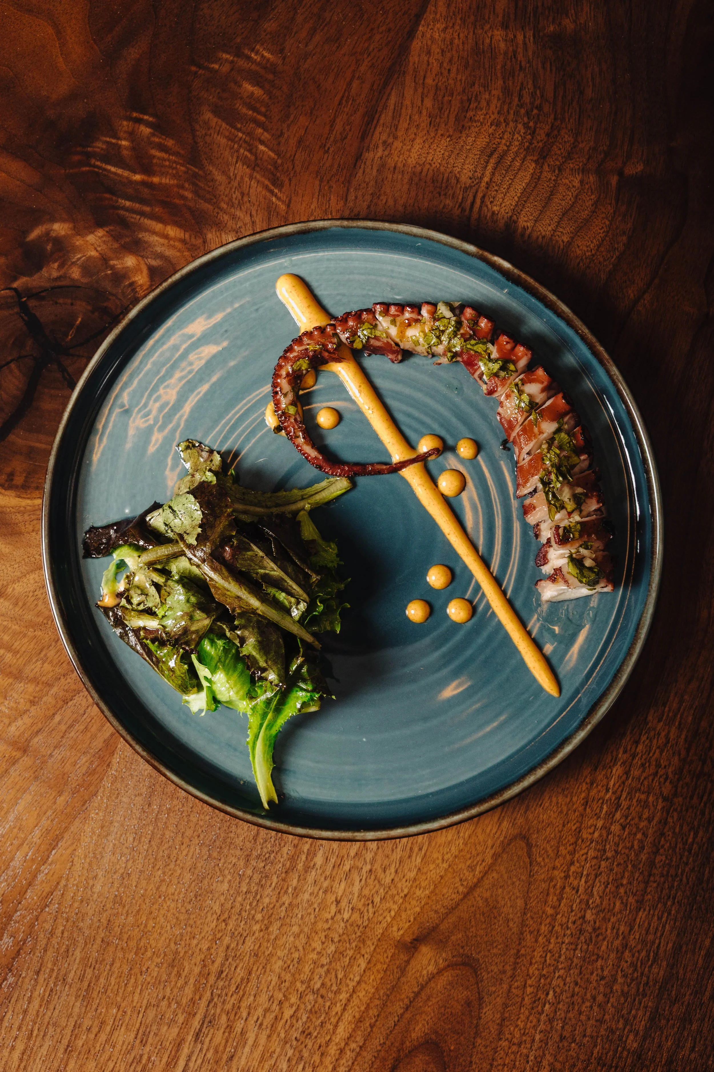 Sliced seared tuna with chopped herbs, a grilled octopus tentacle, a fresh green salad, and mustard sauce on a blue ceramic plate.