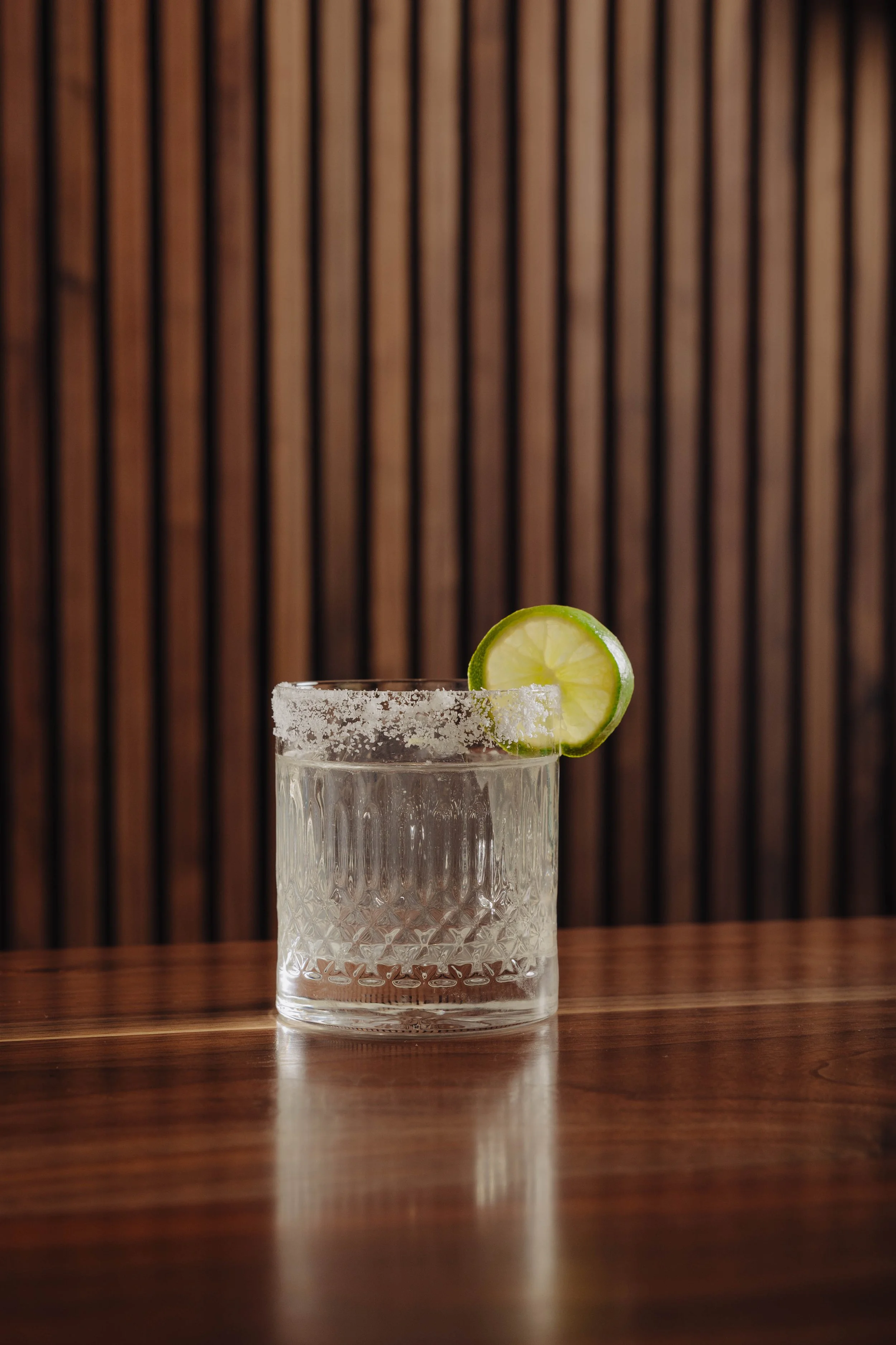 A clear glass containing a drink with a salted rim and garnished with a lime wedge, placed on a wooden surface with a wooden slat background.