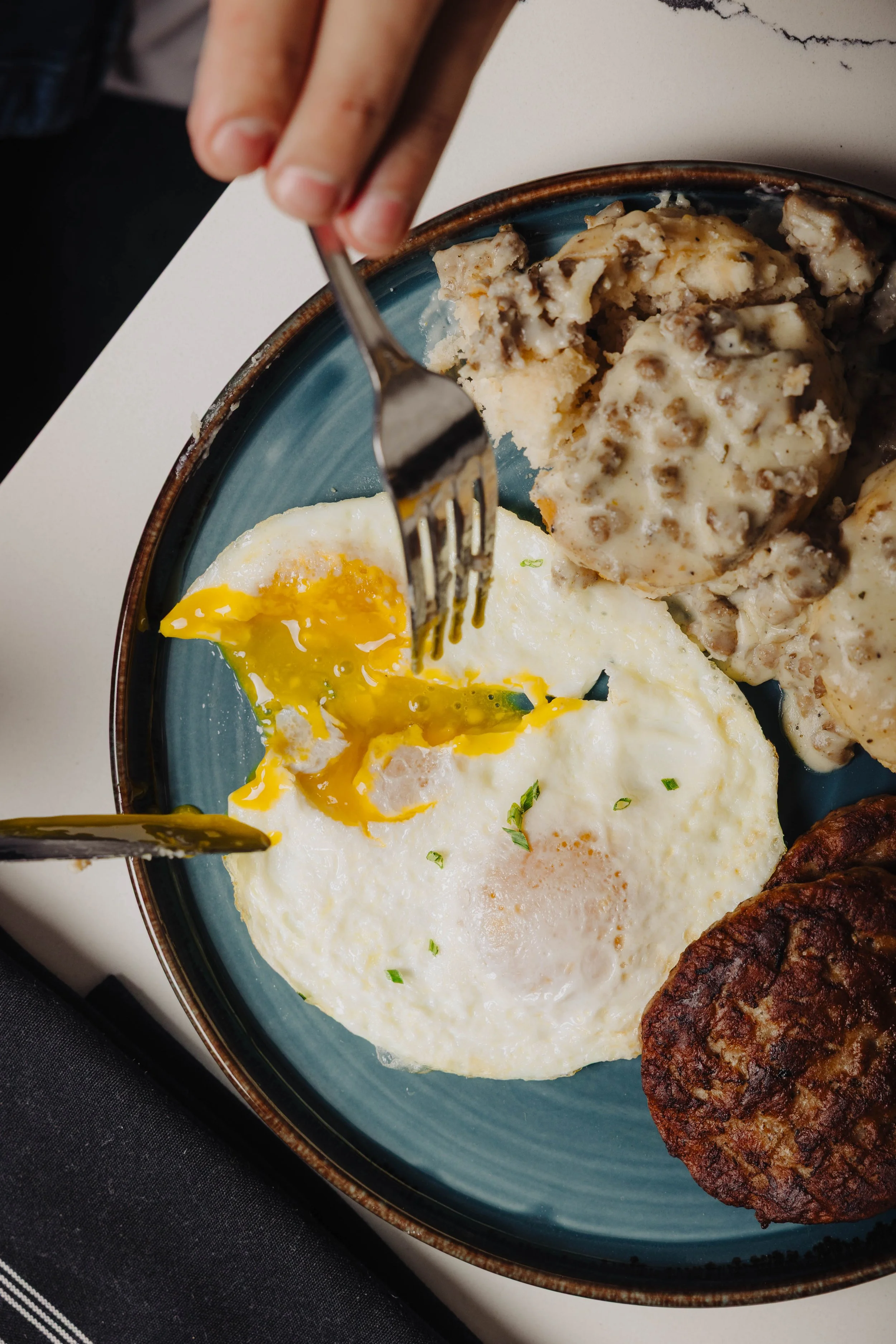 Fried egg with a runny yolk, sausage gravy with biscuits, and a crispy breakfast sausage patty on a blue plate.