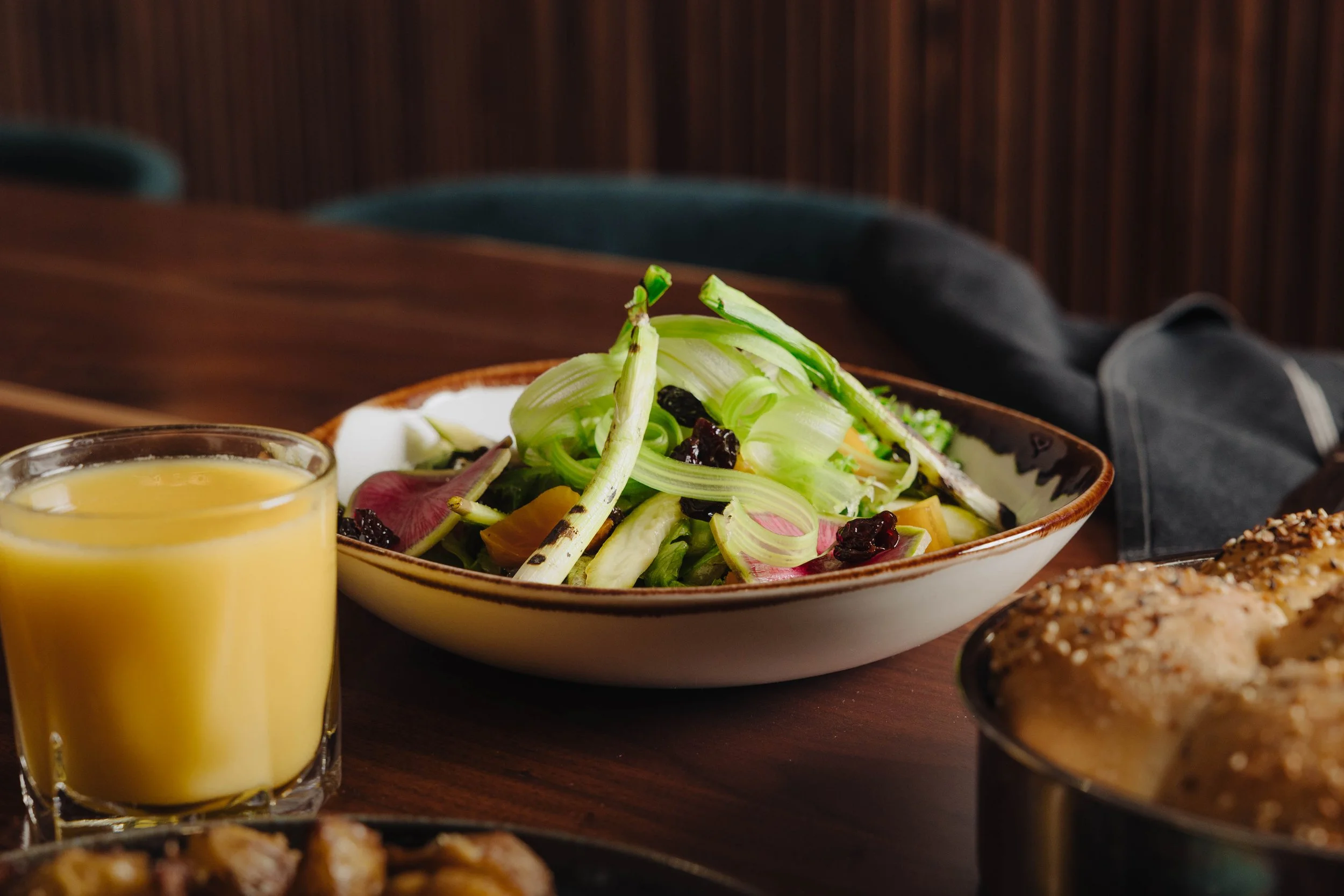 A bowl of salad with sliced vegetables and dried berries, a glass of yellow juice, and a dish of baked food on a wooden table.