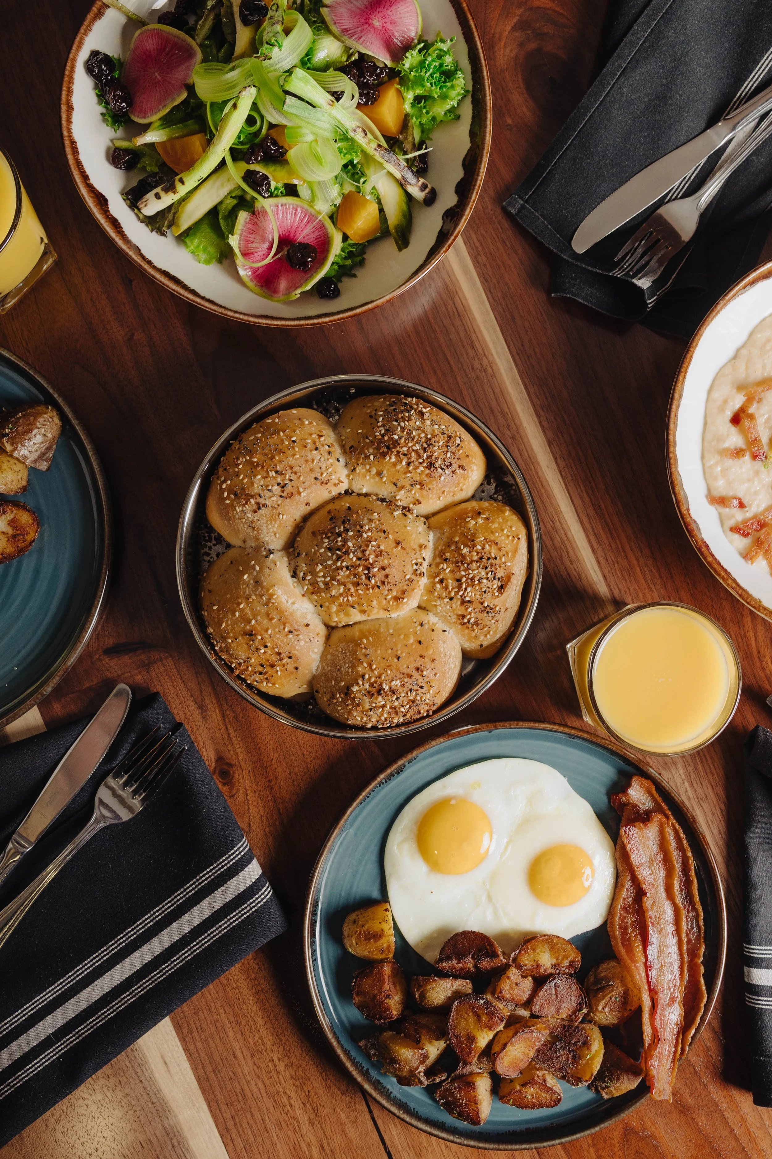 Breakfast spread with fried eggs, bacon, roasted potatoes, dinner rolls, salad with greens, radishes, and dried cranberries, and a glass of orange juice on a wooden table with black napkins and silverware.
