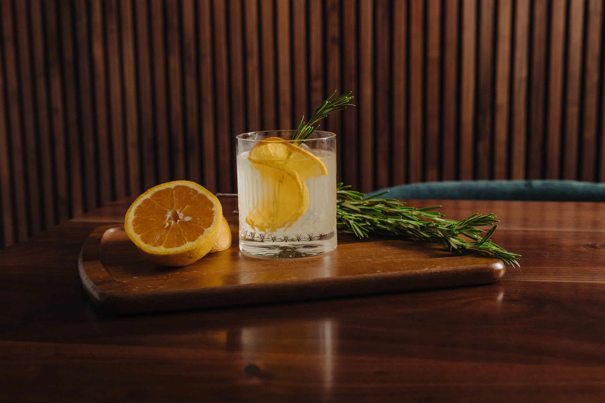 A glass of lemonade with lemon slices and rosemary on a wooden tray, next to a halved lemon, on a wooden table with a wood-paneled background.