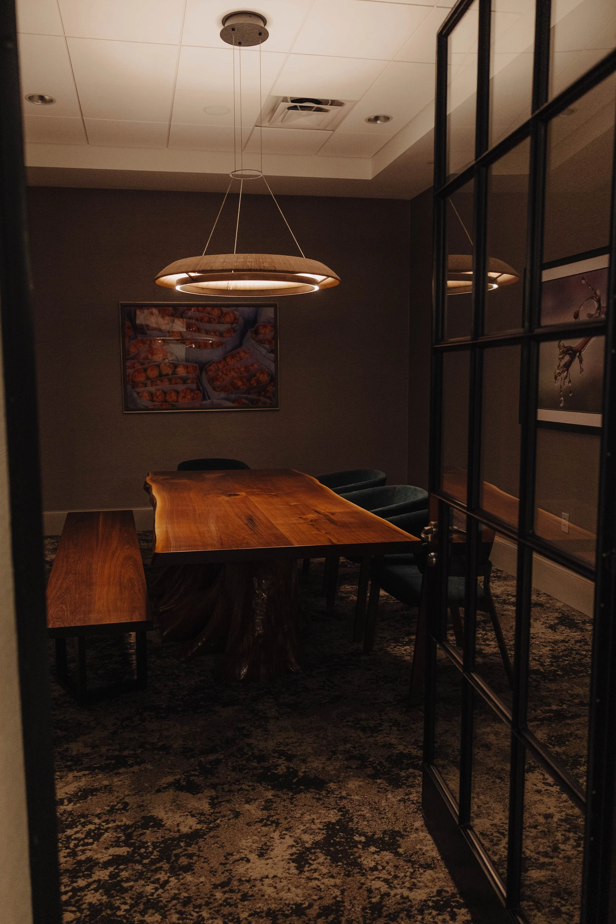 A dimly lit conference room with a wooden table, two chairs, a bench, wall art, and a large circular light fixture hanging from the ceiling.