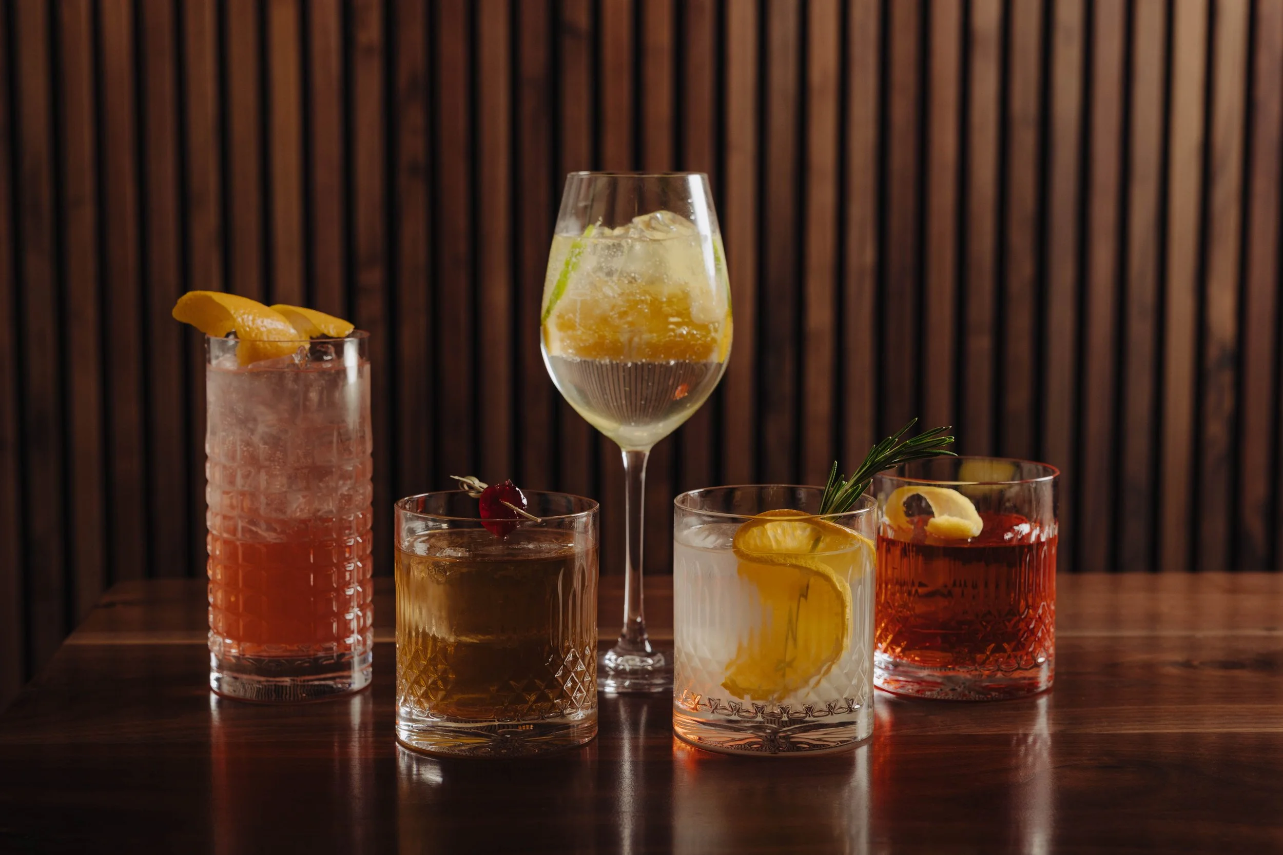 Five colorful cocktails on a wooden table against a wooden panel background.