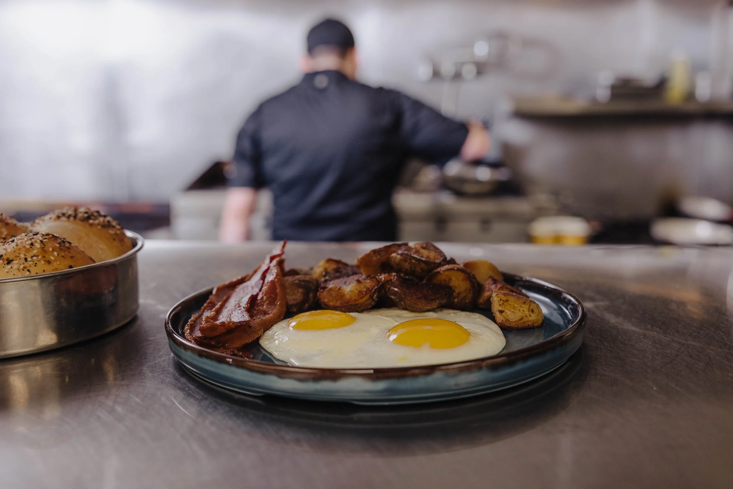 A plate with two sunny-side-up eggs, bacon, and roasted potatoes on a counter in a restaurant kitchen with a chef preparing food in the background.