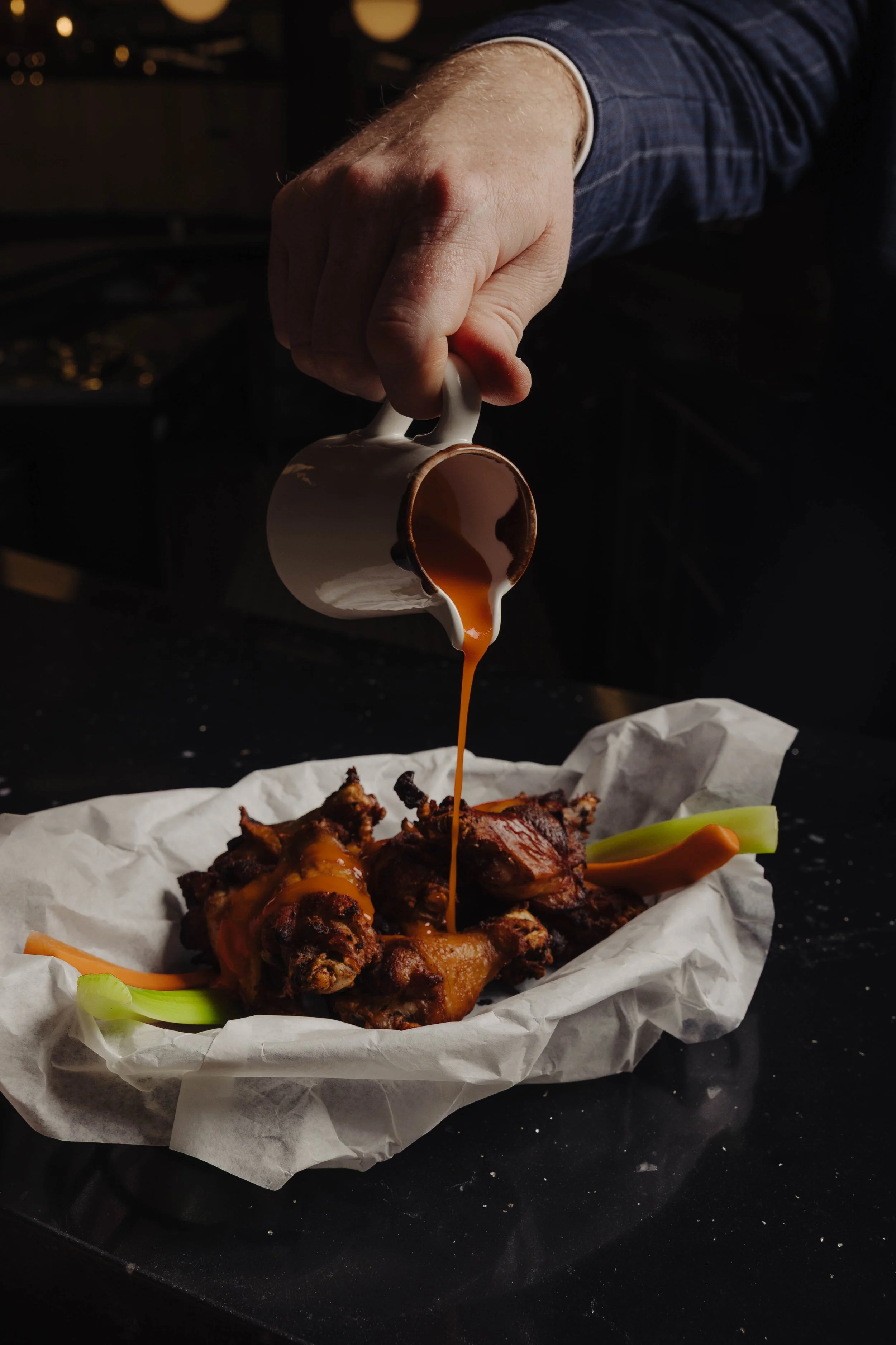 A person in a blue checked shirt pouring brown sauce onto grilled chicken wings and celery sticks on white parchment paper.