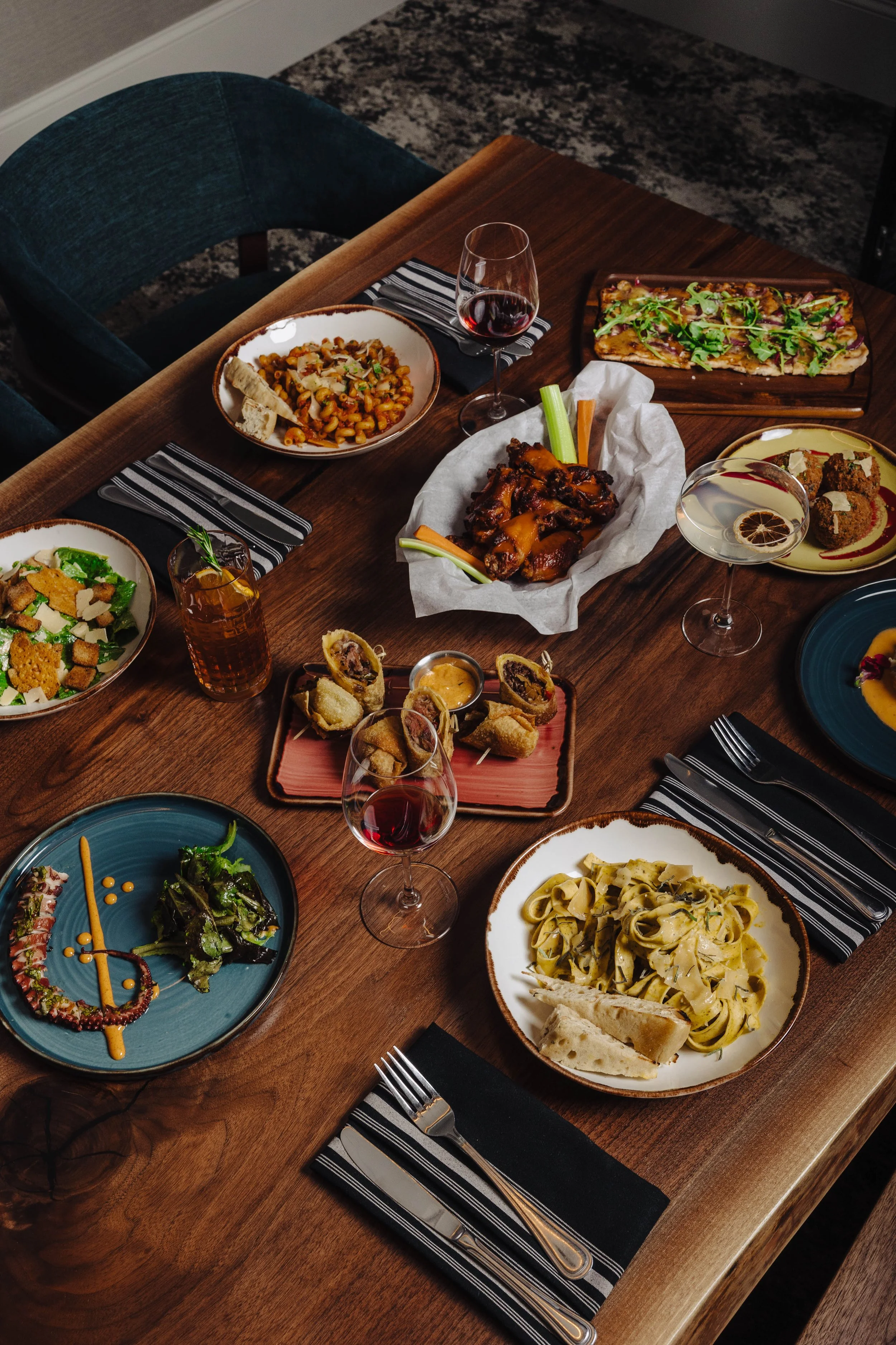 A dinner table with various dishes including pasta, salad, pizza, wings, and desserts, along with glasses of red and white wine, set with black and white striped napkins and silverware.