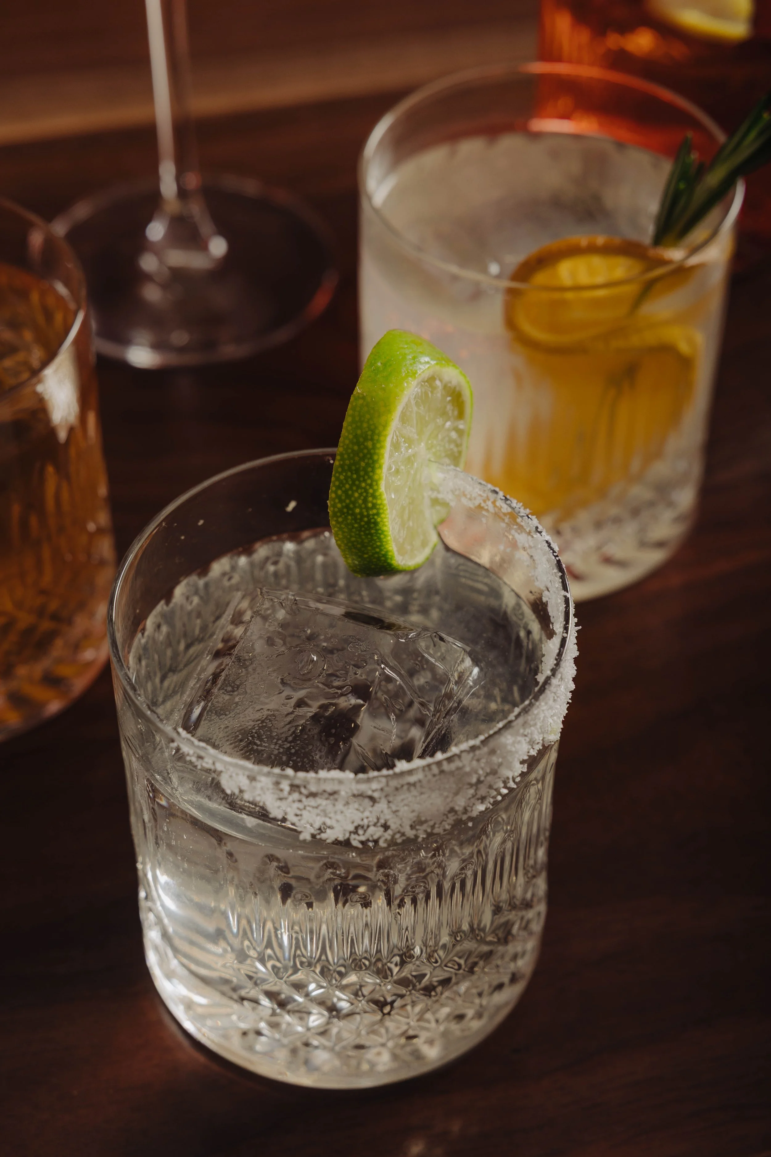 Close-up of a glass of clear cocktail with a salted rim, lime wedge garnish, and ice cubes on a dark wooden table, with other drink glasses in the background.