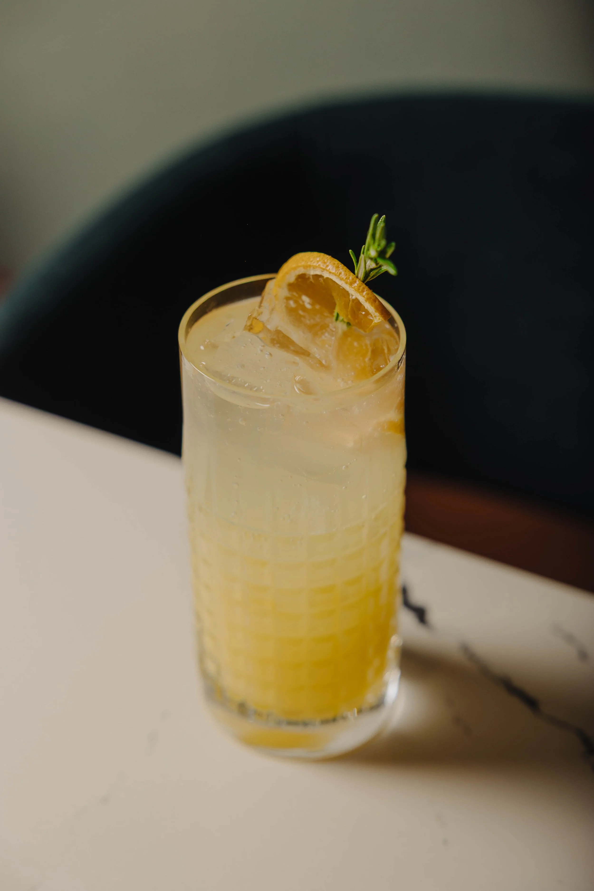 A tall glass of lemonade garnished with a lemon wedge and a sprig of rosemary on top, placed on a white surface.