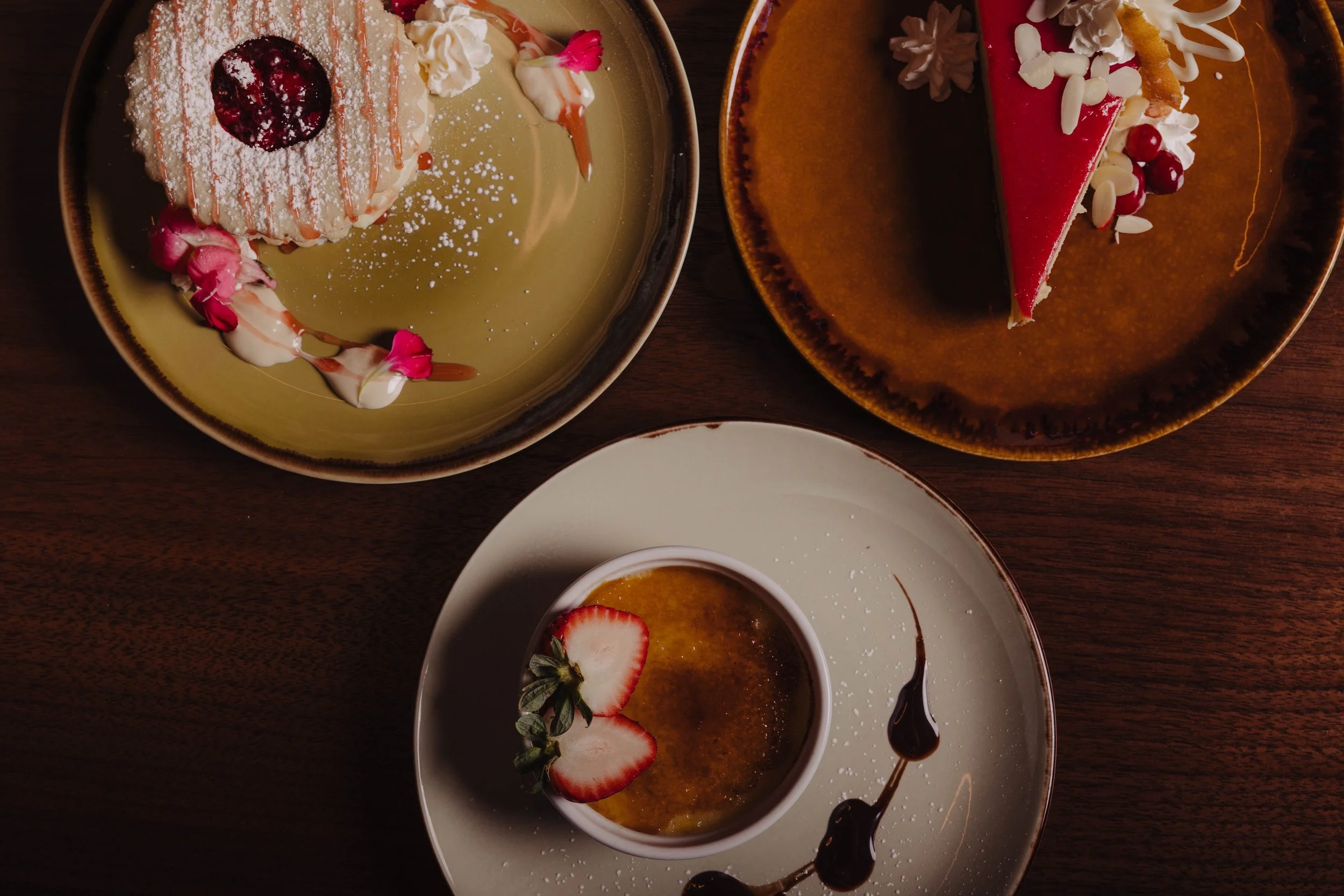 Three plates of dessert on a wooden table, featuring a strawberry tart with a dollop of whipped cream, a red mousse cake with white chocolate shavings, and a crème brûlée garnished with strawberry slices and chocolate drizzle.