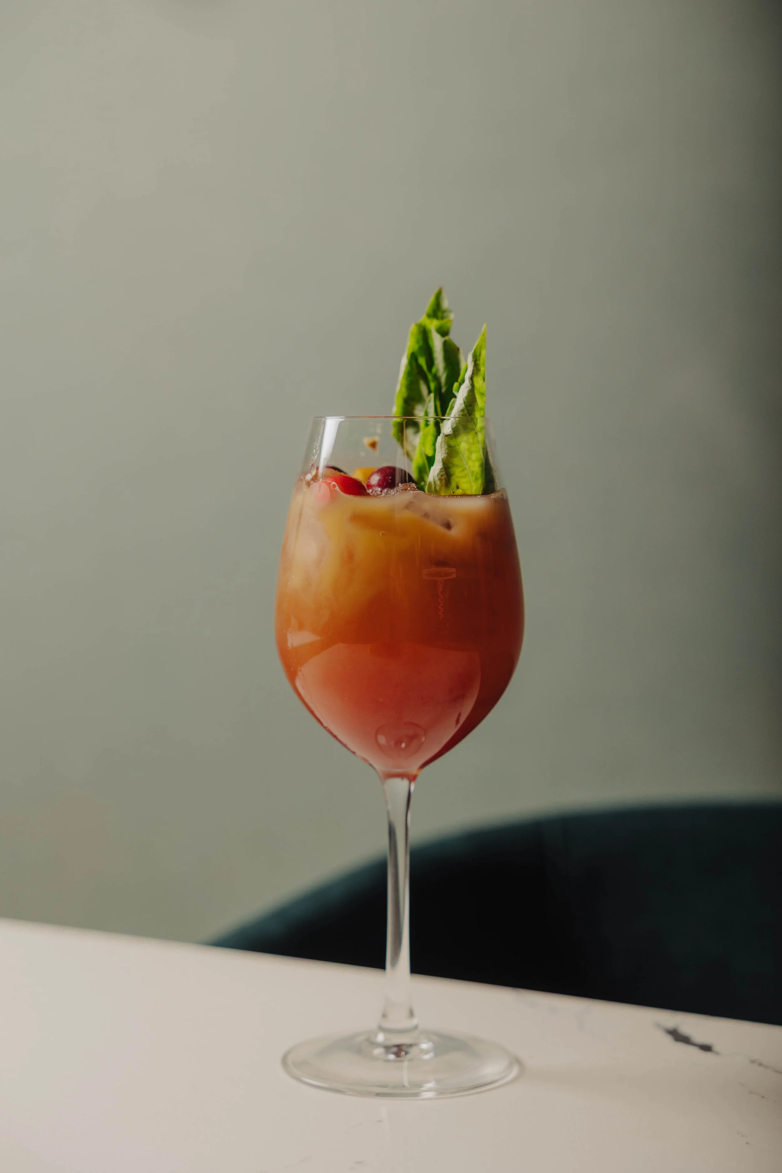 A tall cocktail glass filled with a pinkish-orange drink, garnished with green leafy herbs and red berries.