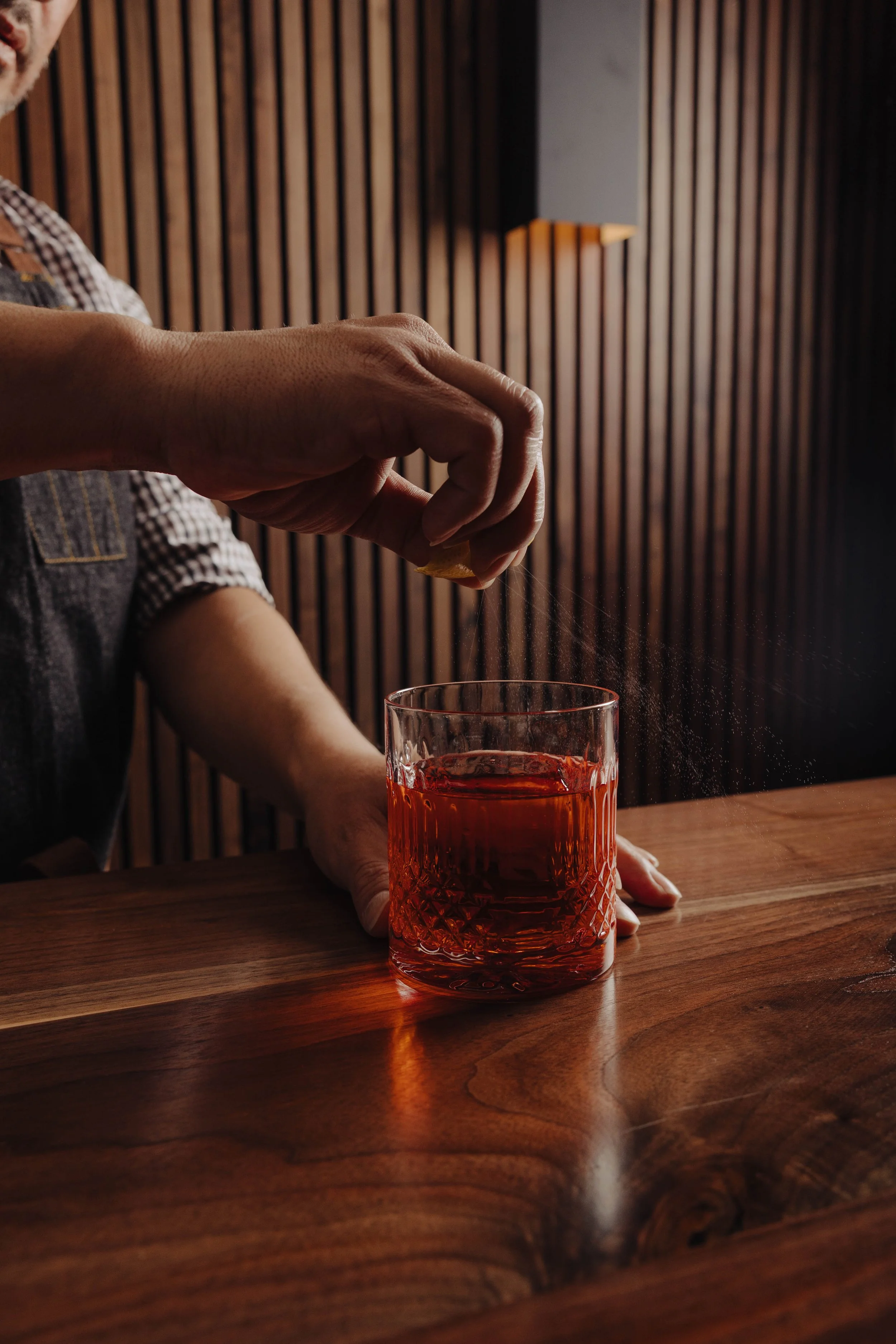 A person sprinkling salt into a glass of red beverage on a wooden table, with a wood-paneled wall in the background.