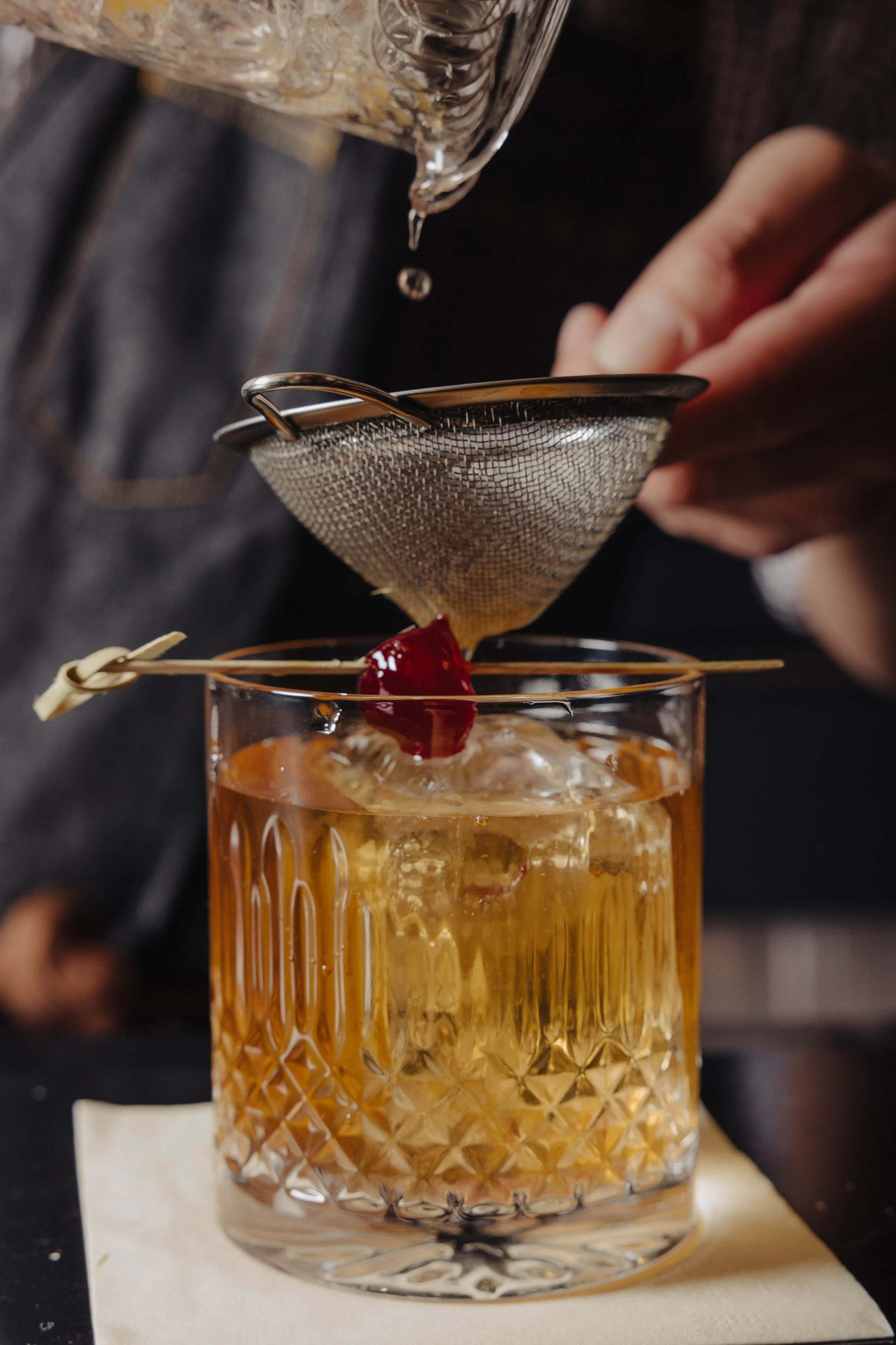 A bartender straining an amber-colored cocktail into a glass with a cherry garnish, using a metal mesh strainer held over the glass.