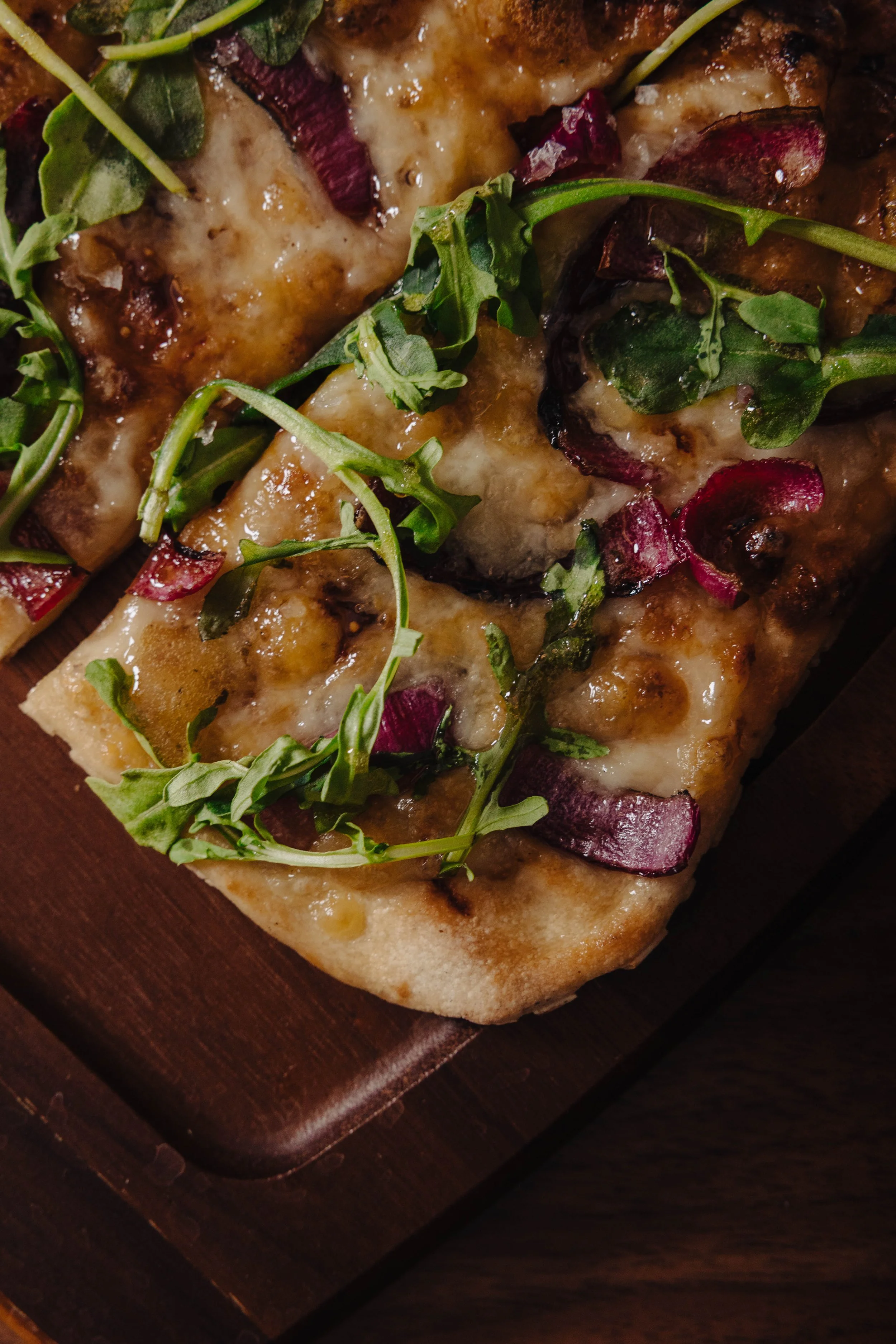 Close-up of a freshly baked flatbread pizza topped with melted cheese, roasted red onions, and fresh arugula leaves on a wooden serving board.