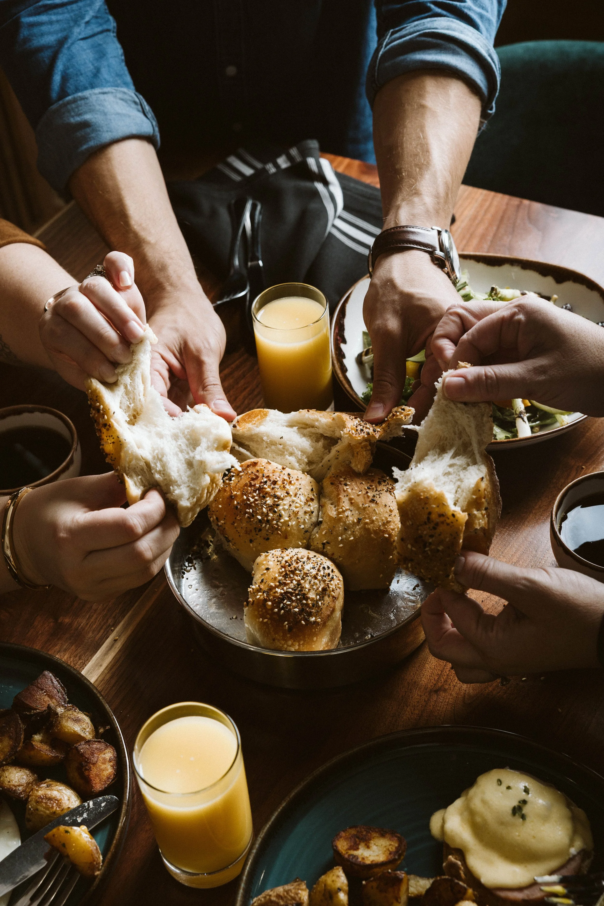 People sharing fresh bread rolls at a breakfast table with orange juice and various dishes.