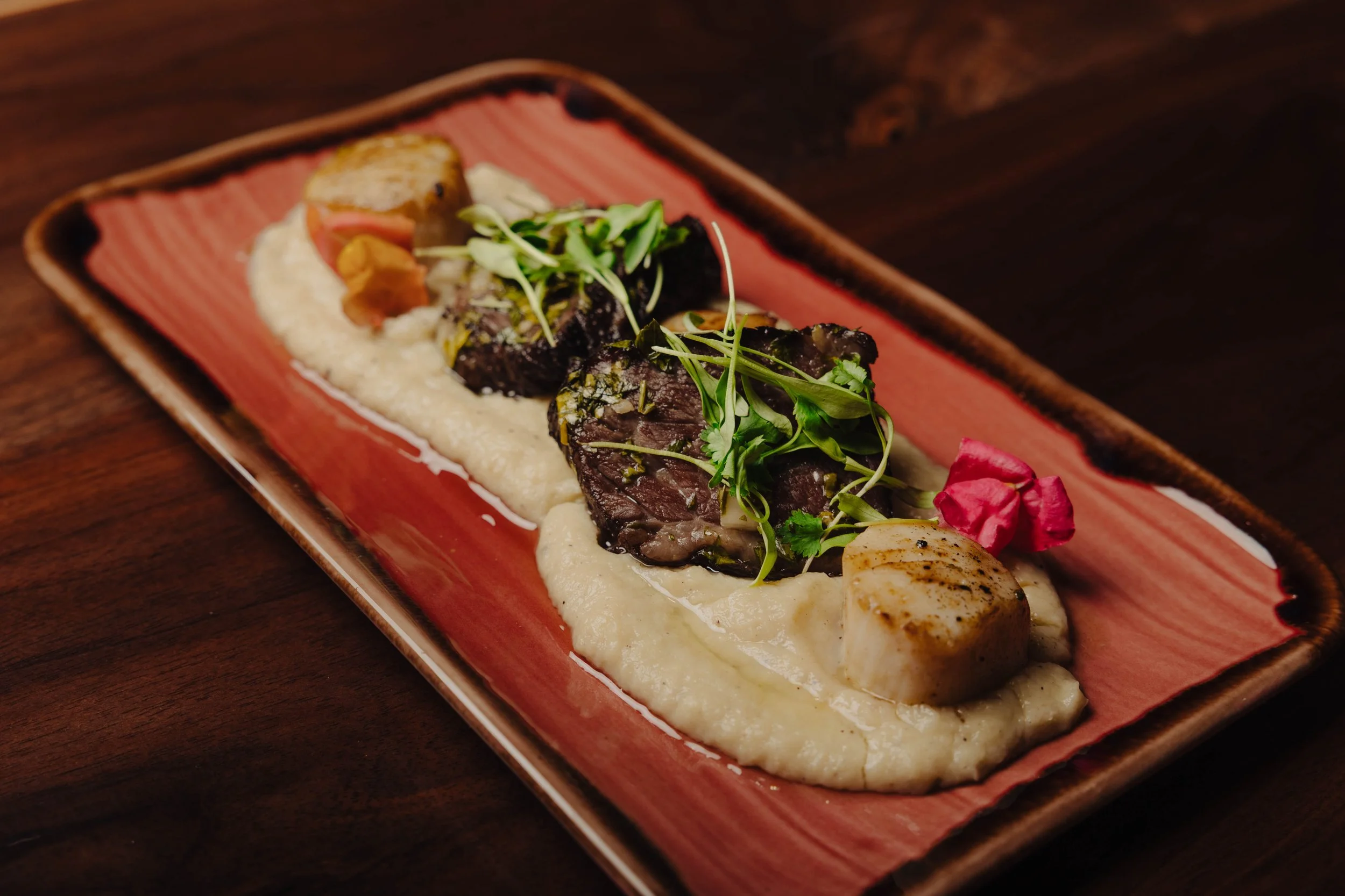 A plated dish featuring seared scallops, crispy short rib, chimichurri, cauliflower smash.