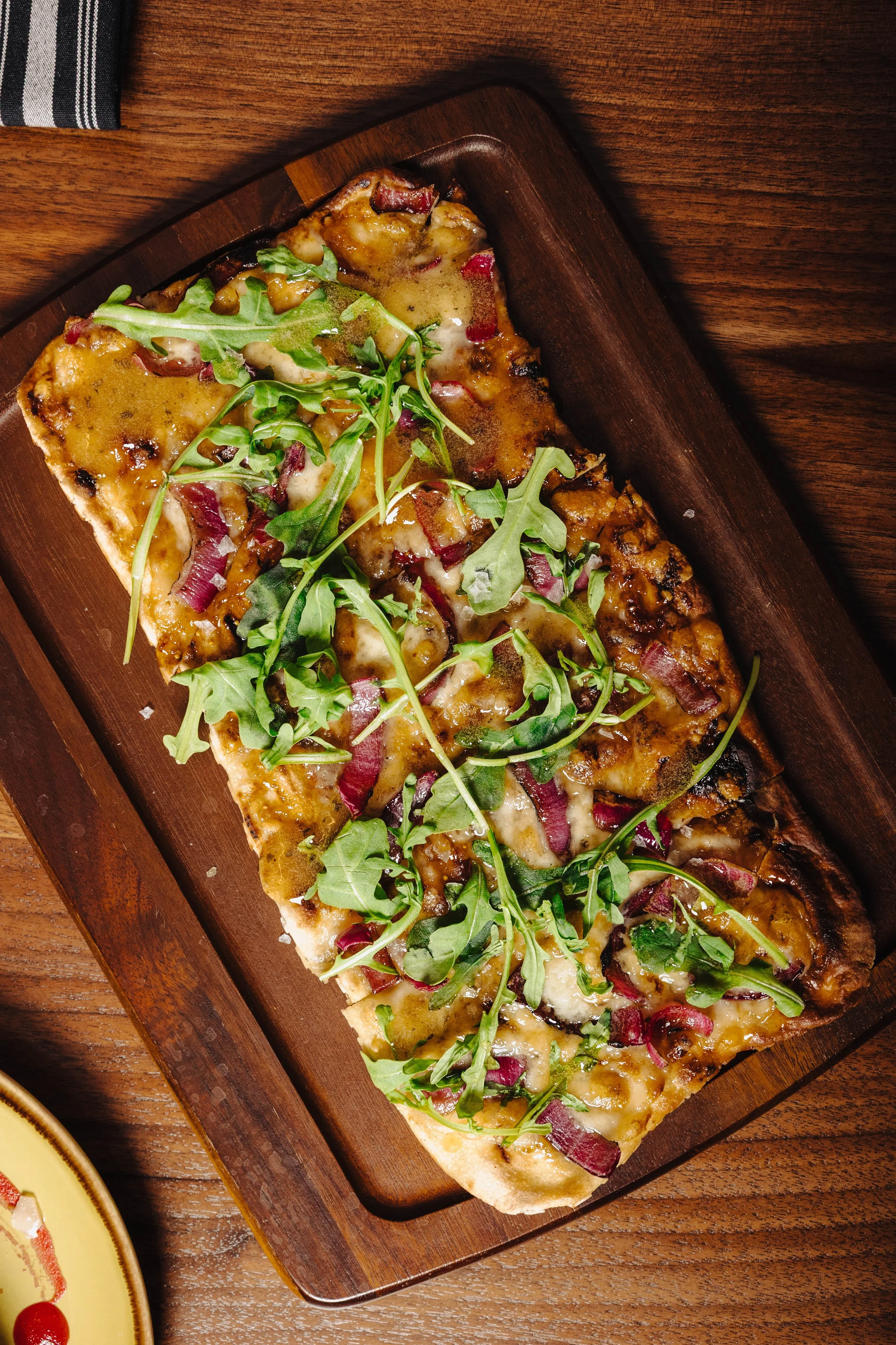 A flatbread pizza topped with cheese, red onions, and fresh arugula leaves on a wooden serving board.