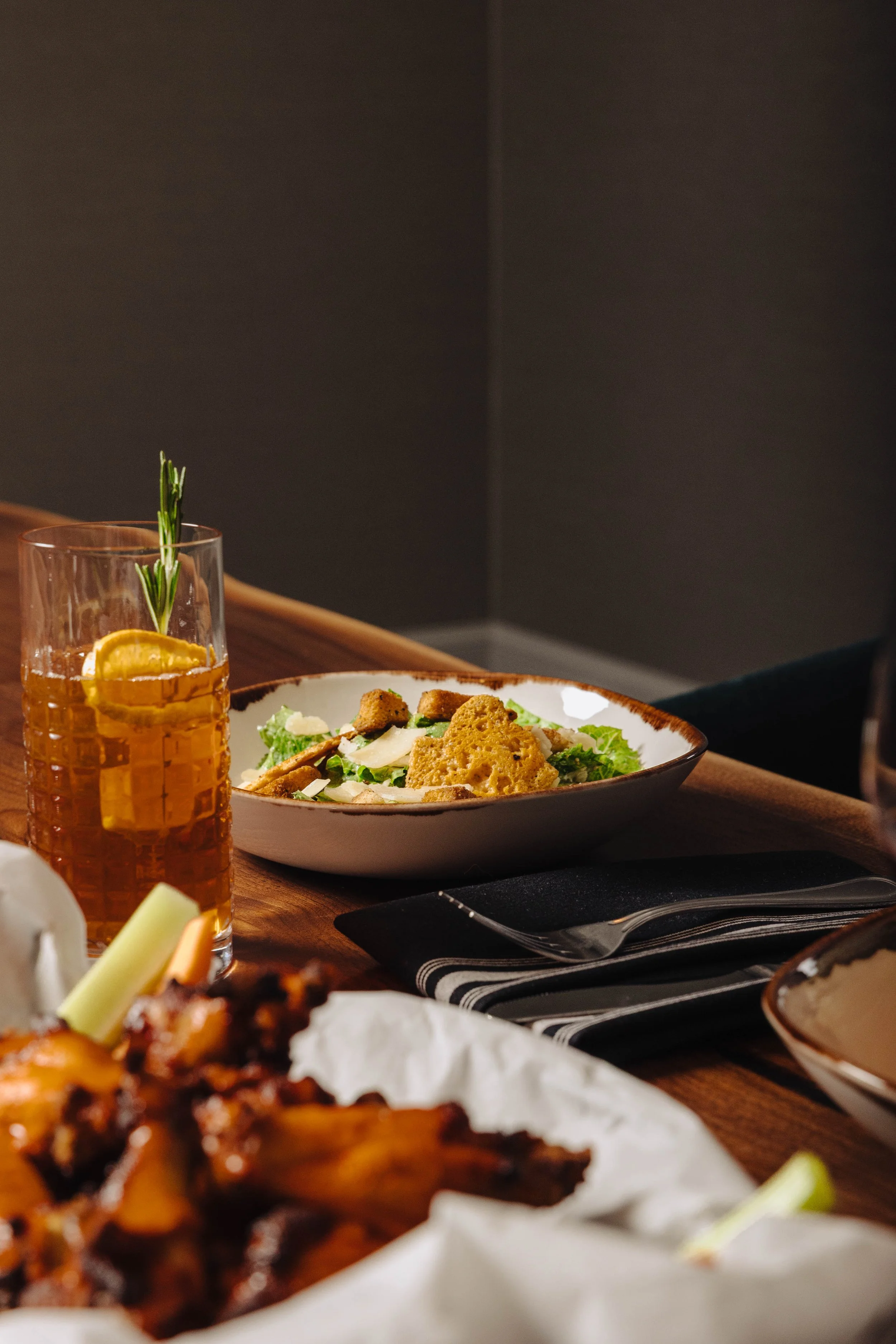 A dining table with a cocktail, a salad, and a plate of ribs on a paper-lined tray, set against a dark background.