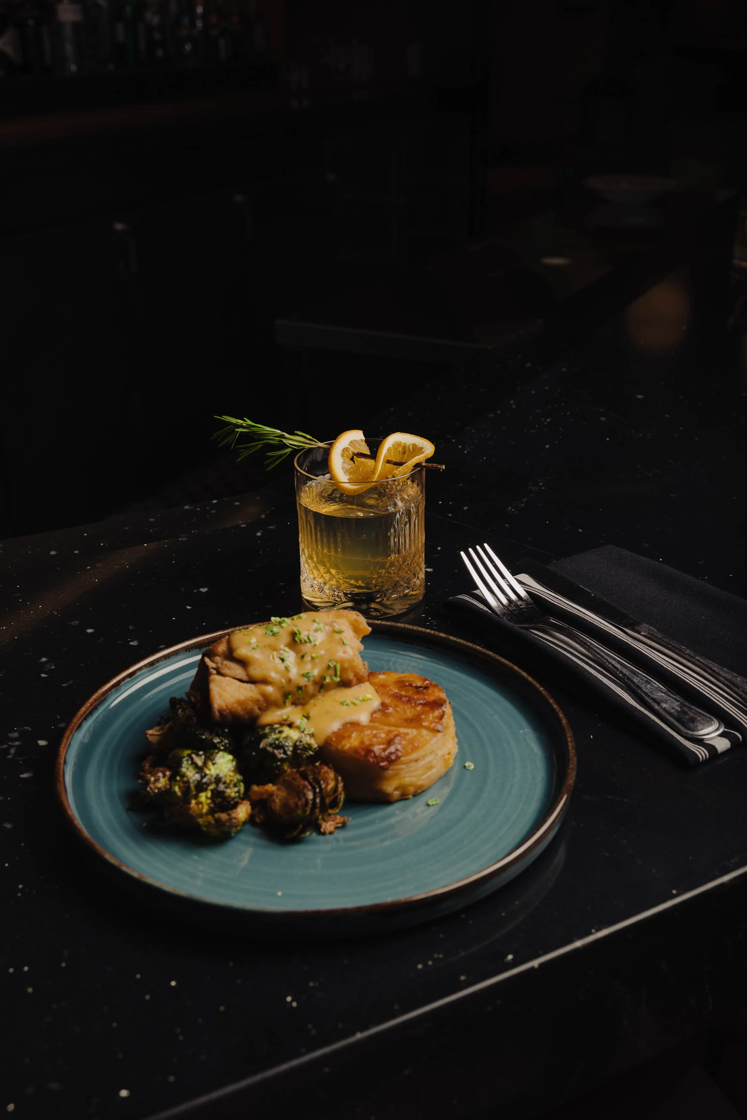 Cornmeal Crusted Lake Trout featuring crusted lake trout, cajun cream sauce, cheesy potatoes, crispy sea salt Brussels. Accompanying the meal is a glass of drink garnished with citrus slices and a sprig of rosemary.