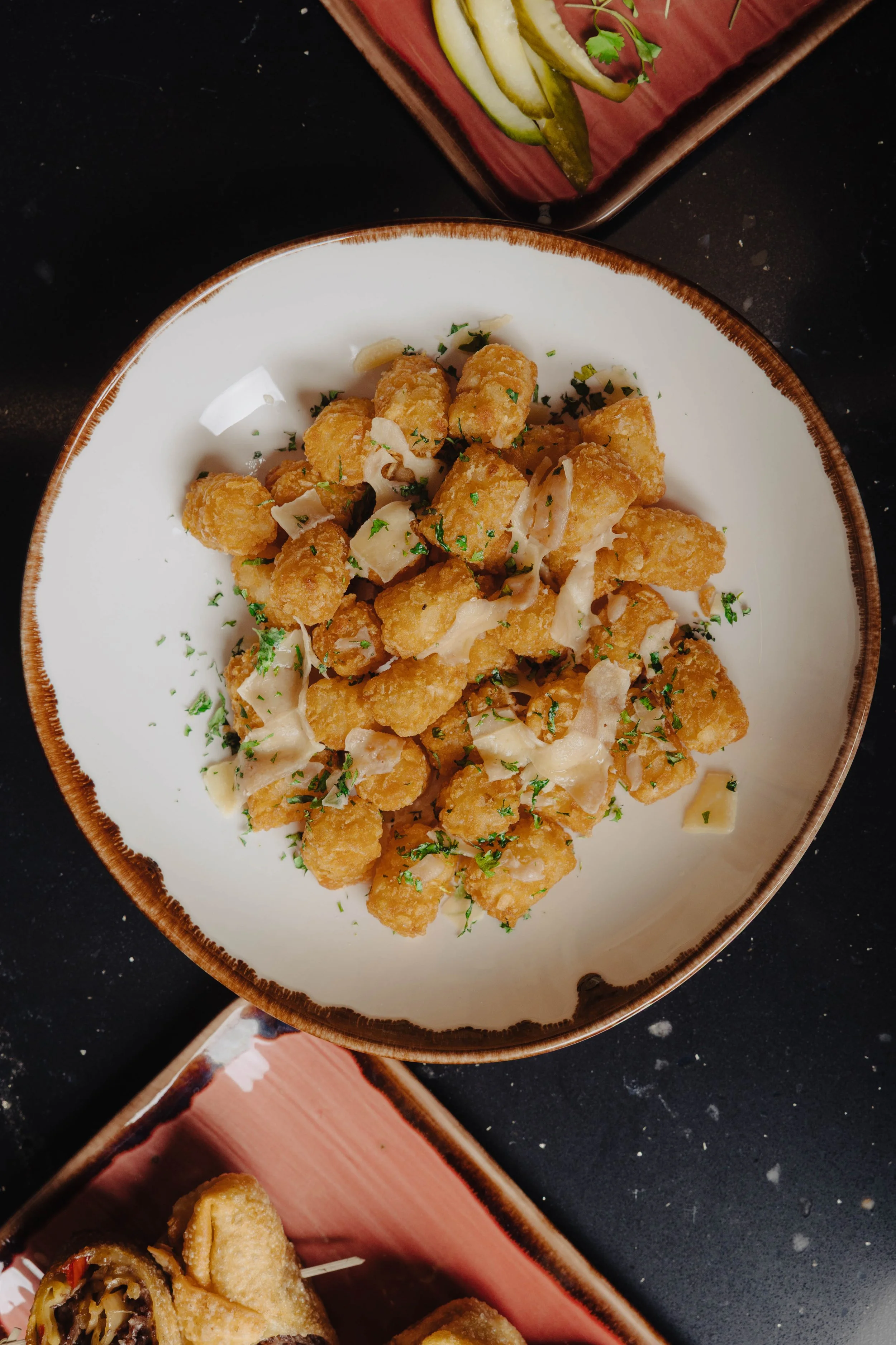 A white bowl with a brown rim filled with fried gnocchi garnished with chopped herbs and cheese shavings, placed on a black surface.