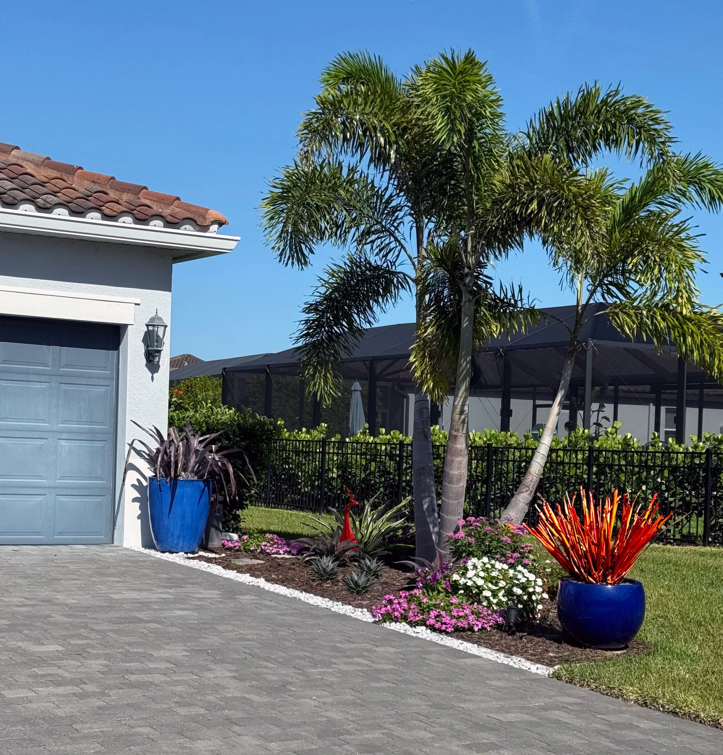 A residential front yard features potted plants, flower beds, a tree, and a stone pathway. There is a blue pot with a plant, colorful flowers, and a white fence in the background, with a house to the left and blue sky overhead.