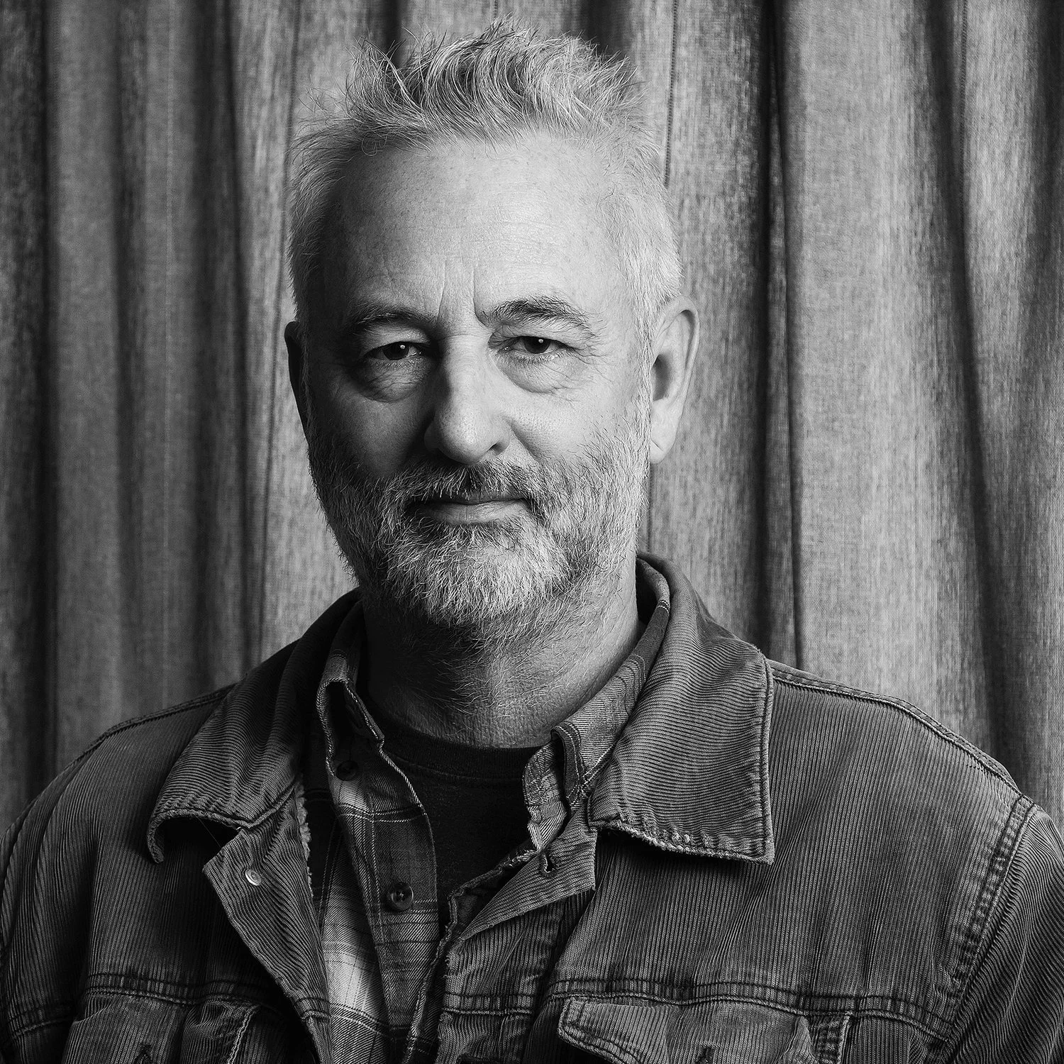 Black and white portrait of a middle-aged man with a beard and mustache, wearing a denim jacket over a plaid shirt, standing in front of a textured curtain.