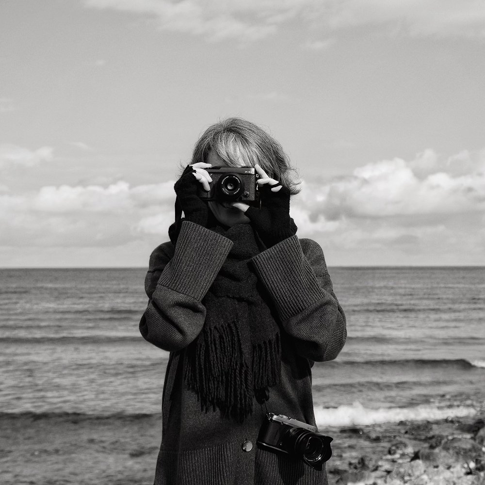 A person stands on a beach holding a camera up to their face, taking a photo, with another camera hanging from their neck. They are dressed warmly with a scarf, sweater, and gloves, and the ocean and sky are in the background.