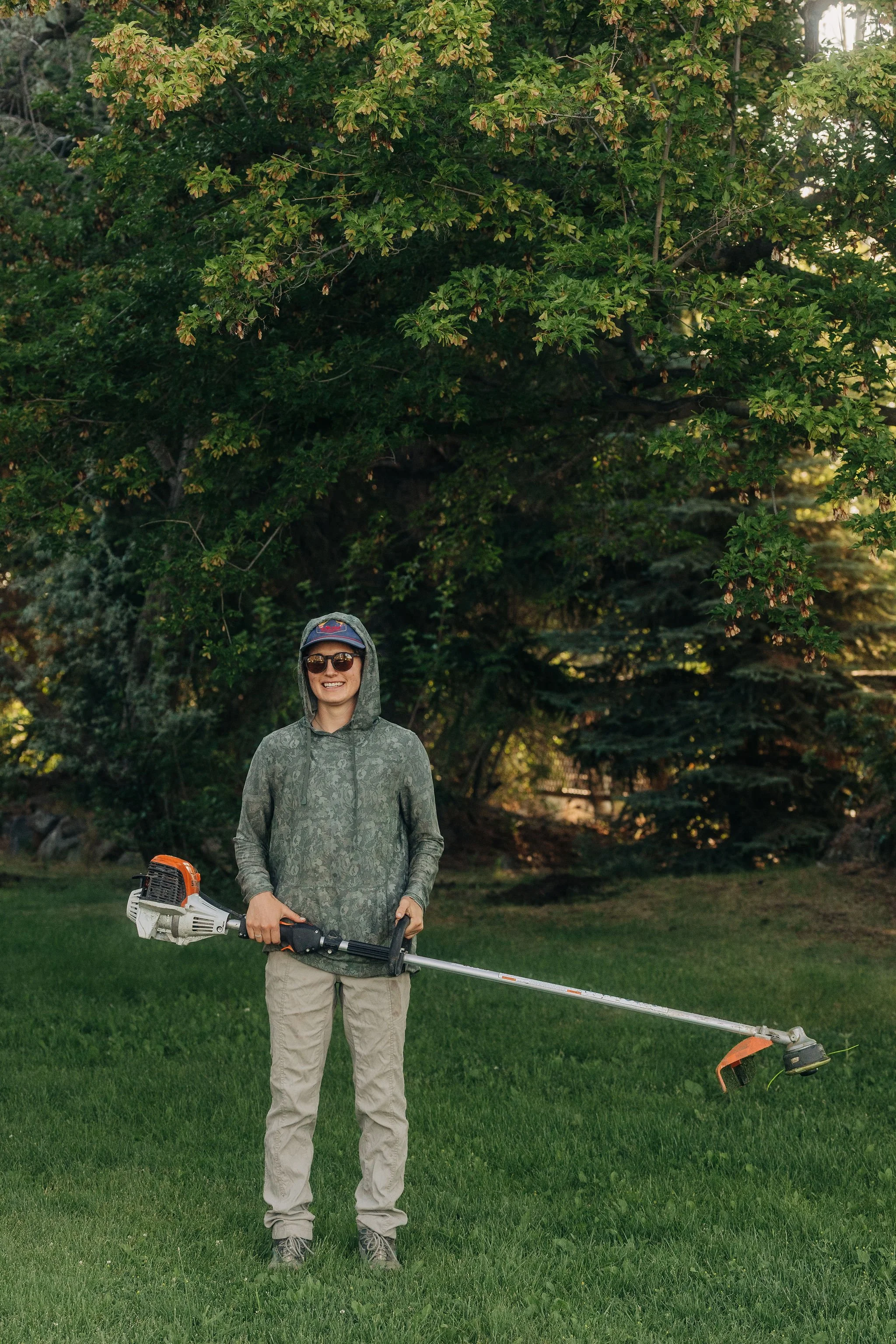 A smiling person wearing sunglasses, a hat, and a gray camouflage hoodie, standing on green grass in a park-like setting with large trees in the background, holding a gas-powered weed trimmer.