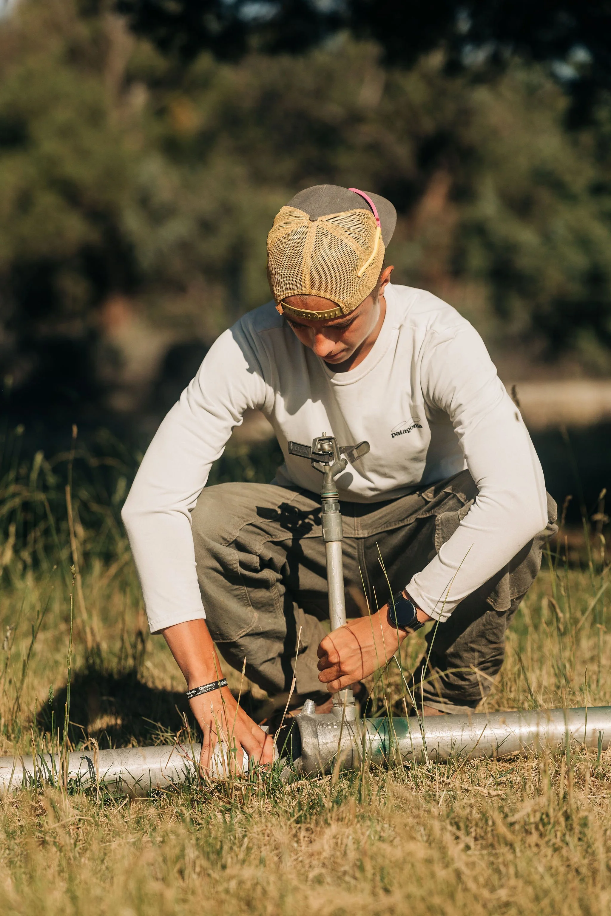 A man wearing a beige cap, sunglasses on top, and a white long-sleeve shirt is kneeling outdoors in a grassy area with trees in the background. He appears to be working on a pipe or piece of equipment on the ground.