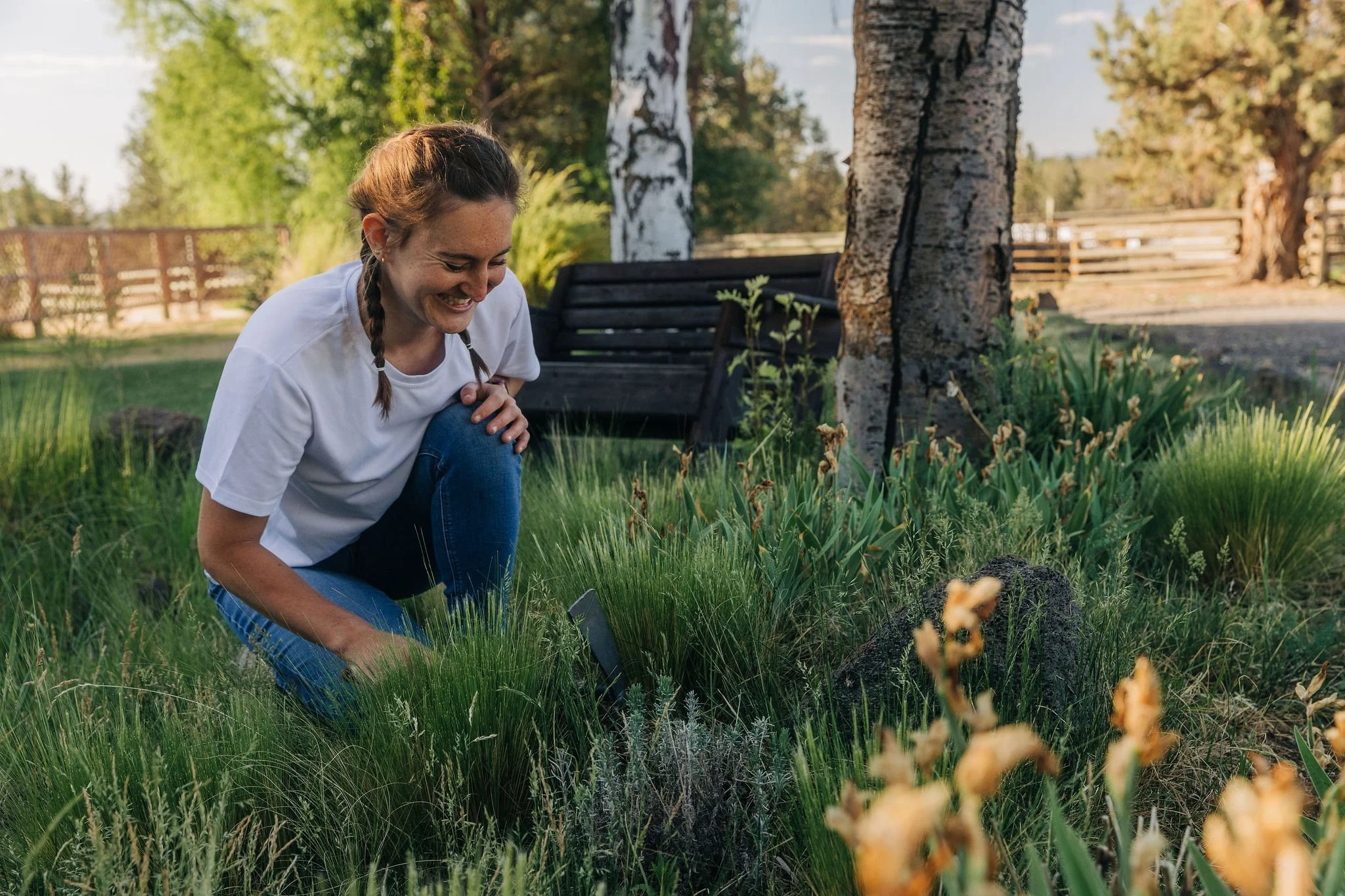 A woman with brown hair in braids, wearing a white t-shirt, crouches in a garden, smiling as she looks at plants. There are trees, a wooden bench, and a wooden fence in the background.