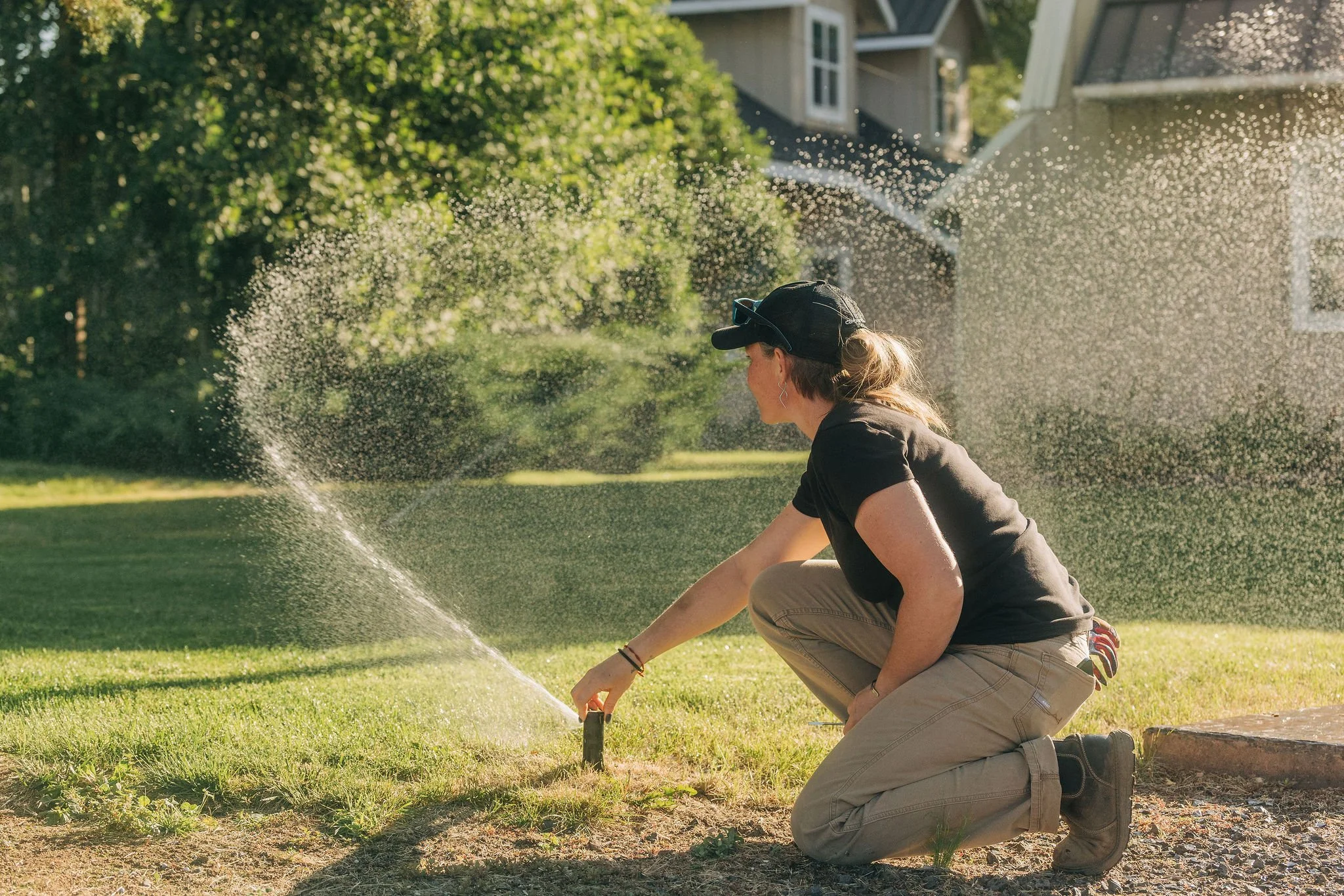 A woman kneeling on the grass near a sprinkler system in a backyard, watering the lawn on a sunny day.