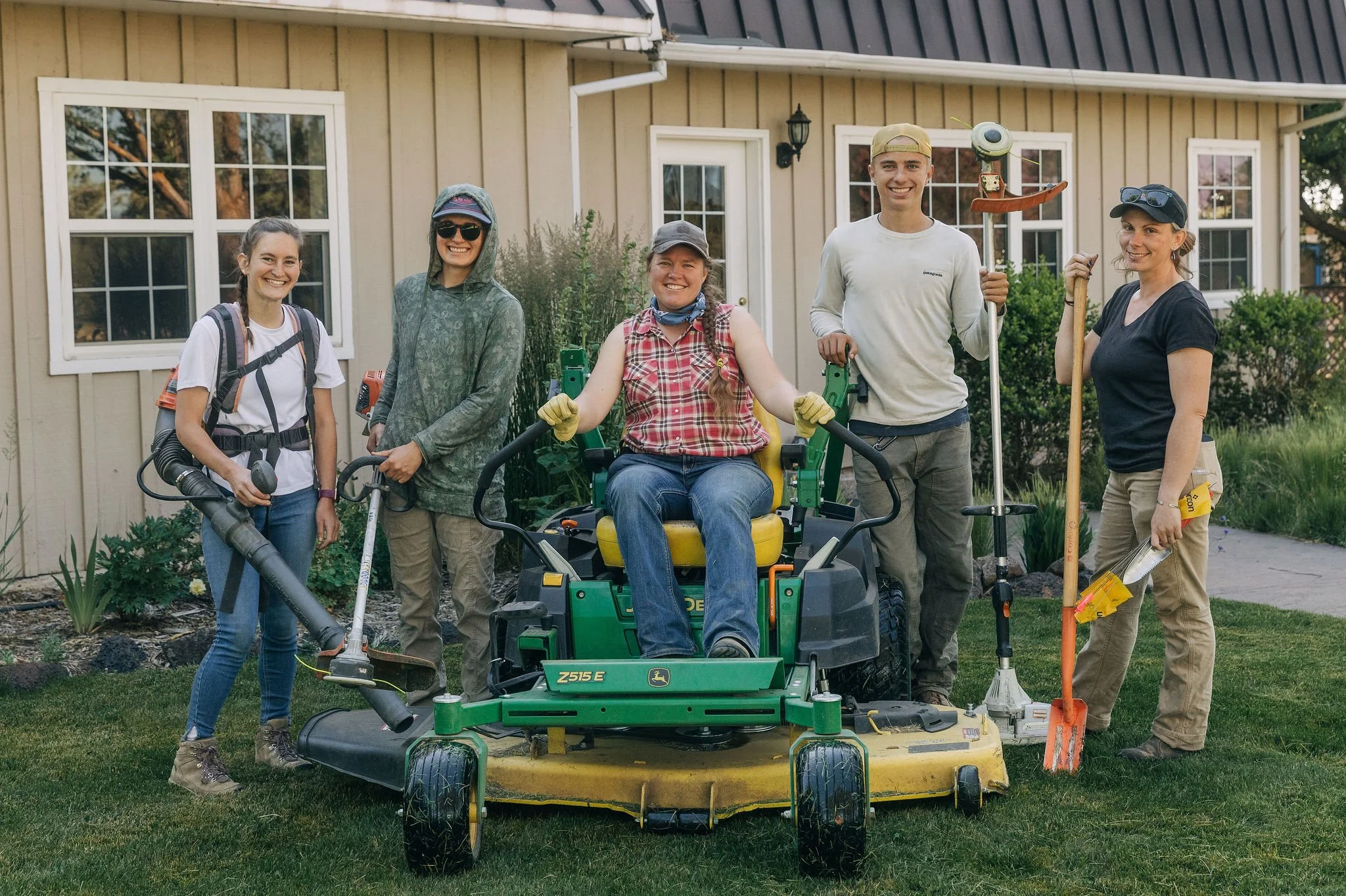Group of five people standing outdoors in front of a house, one person seated on lawn mower, all smiling, holding gardening tools, with garden and shrubs in the background.