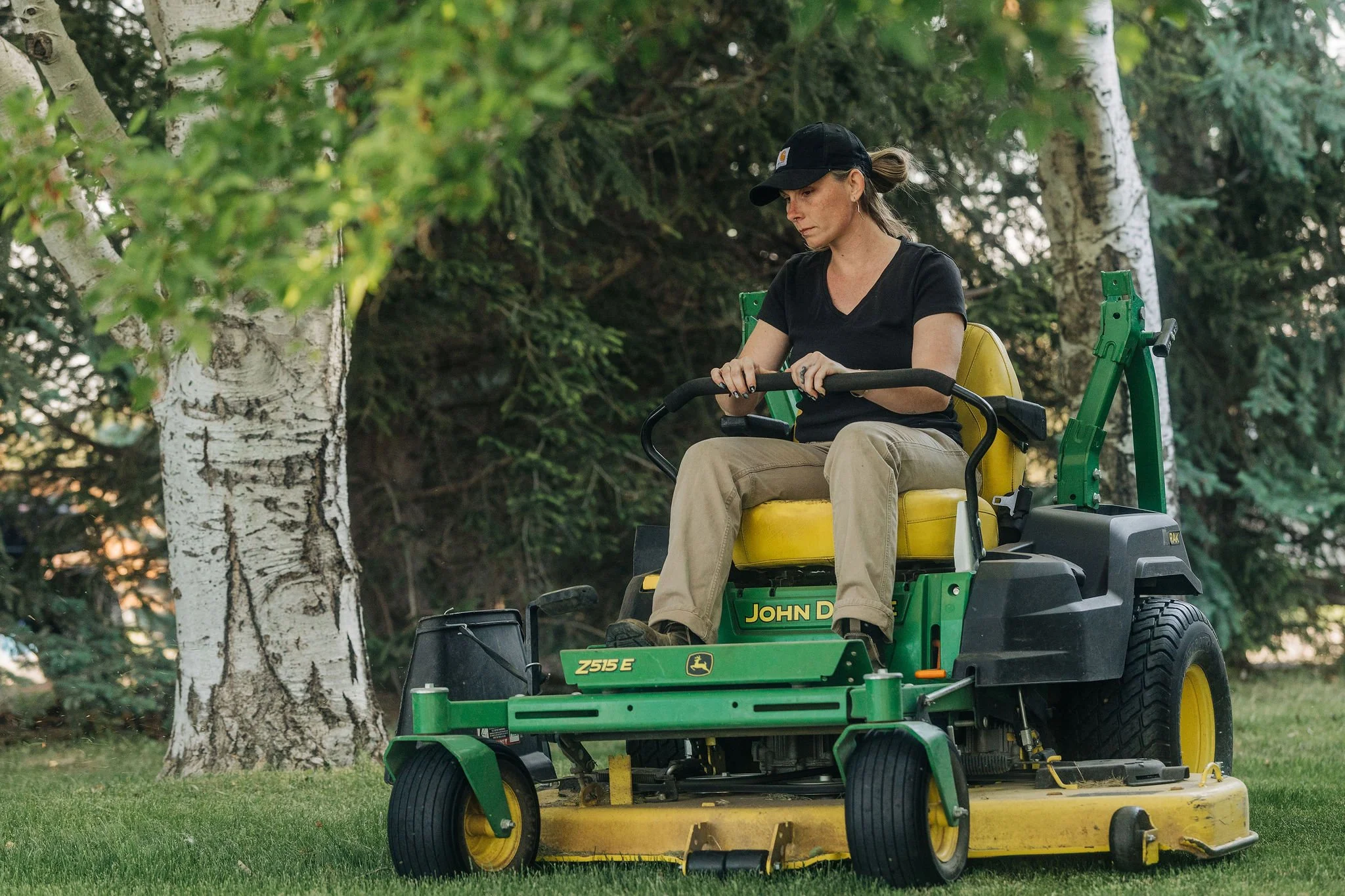 A woman operating a John Deere Z515E riding lawn mower on a grassy yard surrounded by trees, wearing a black t-shirt, beige pants, a black cap, and earrings.