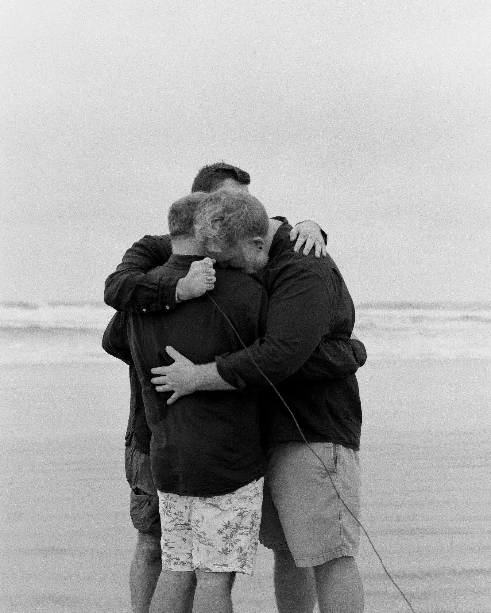 Three men embrace and hug on the beach with ocean waves in the background, one man has his eyes closed and head resting on the other's shoulder.