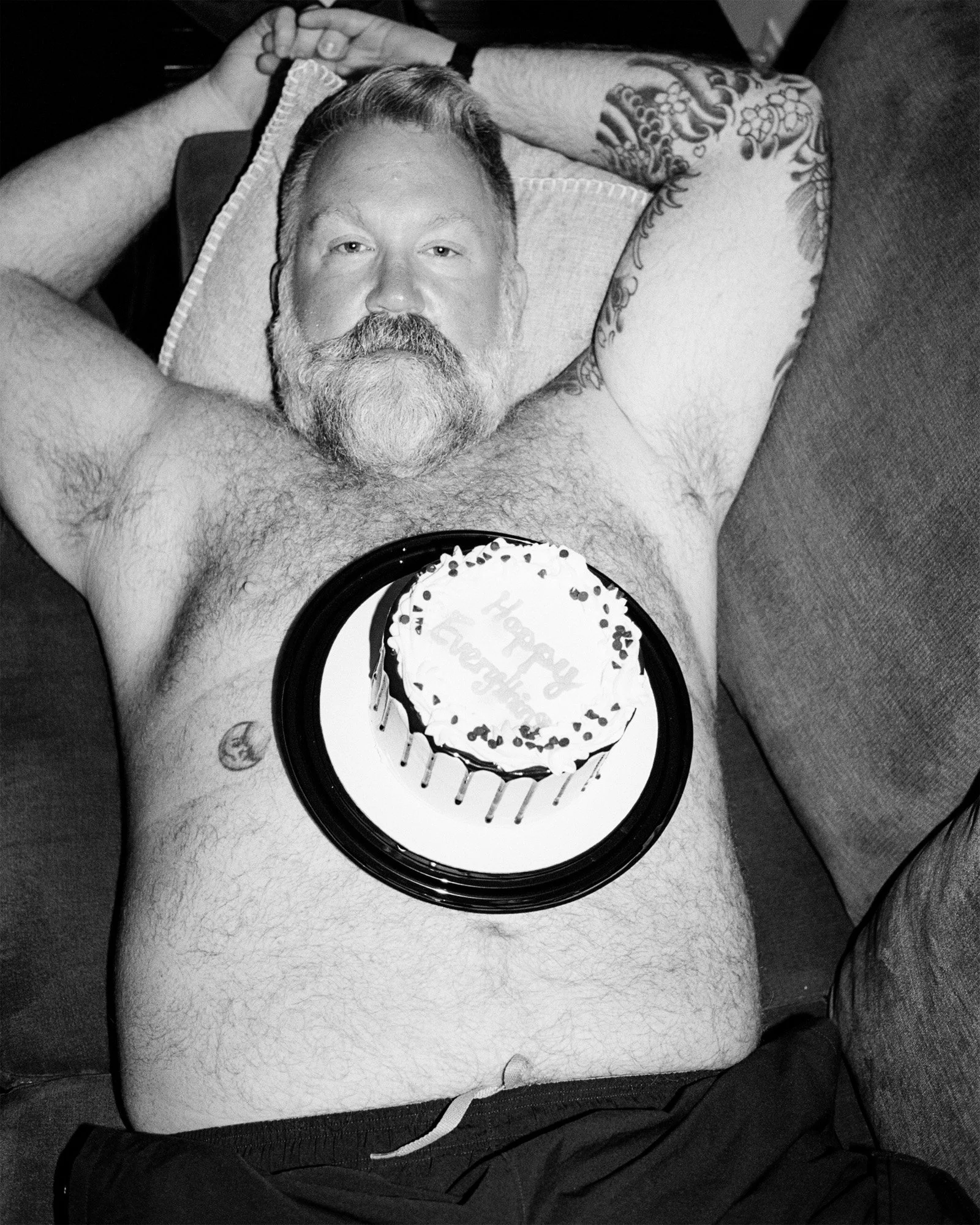 A shirtless man with a beard and tattoos lying on a couch with his arms behind his head, holding a birthday cake with 'Happy Birthday' written on it on his chest.
