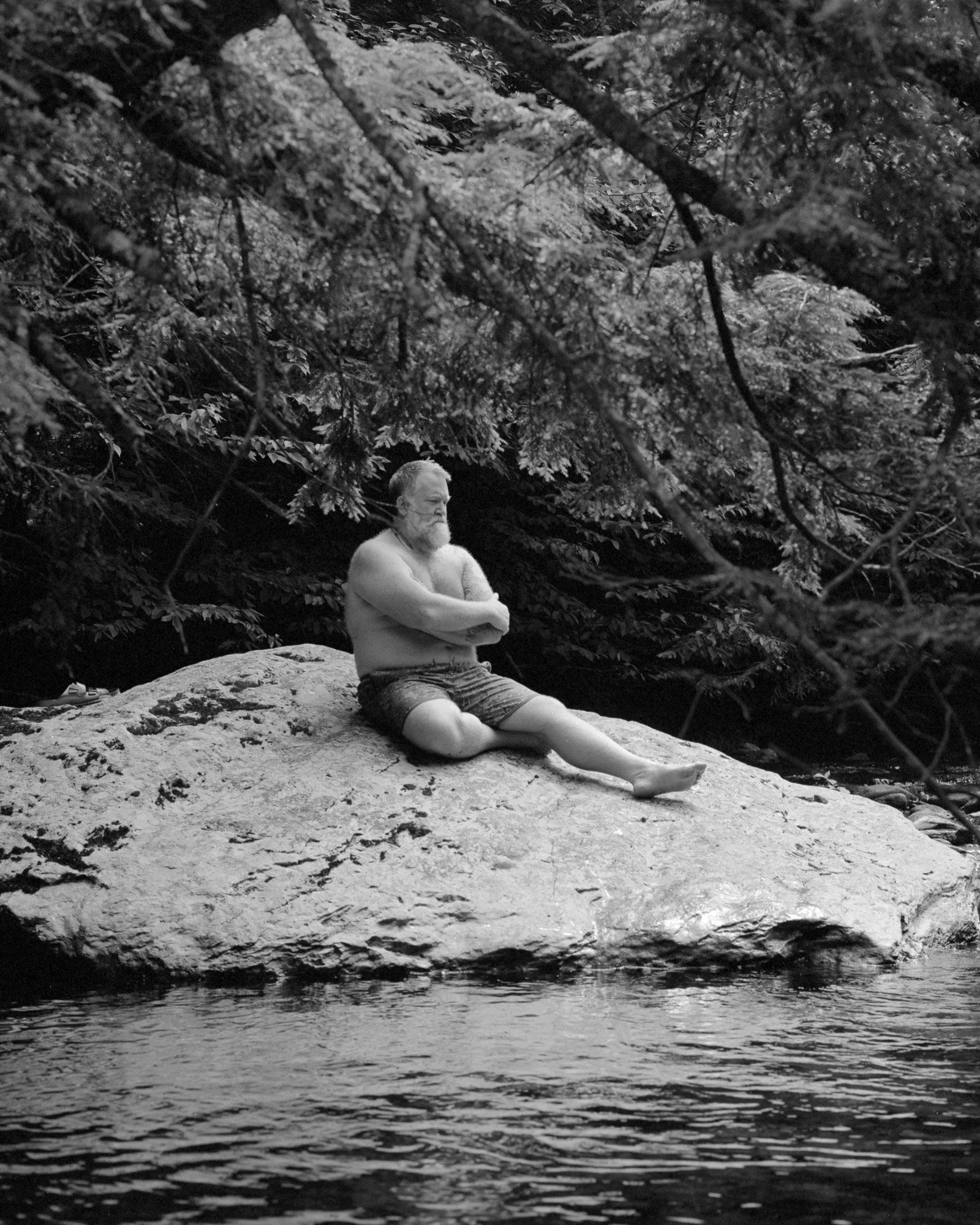 A shirtless elderly man with a beard sitting cross-legged on a large rock near a river, surrounded by trees.