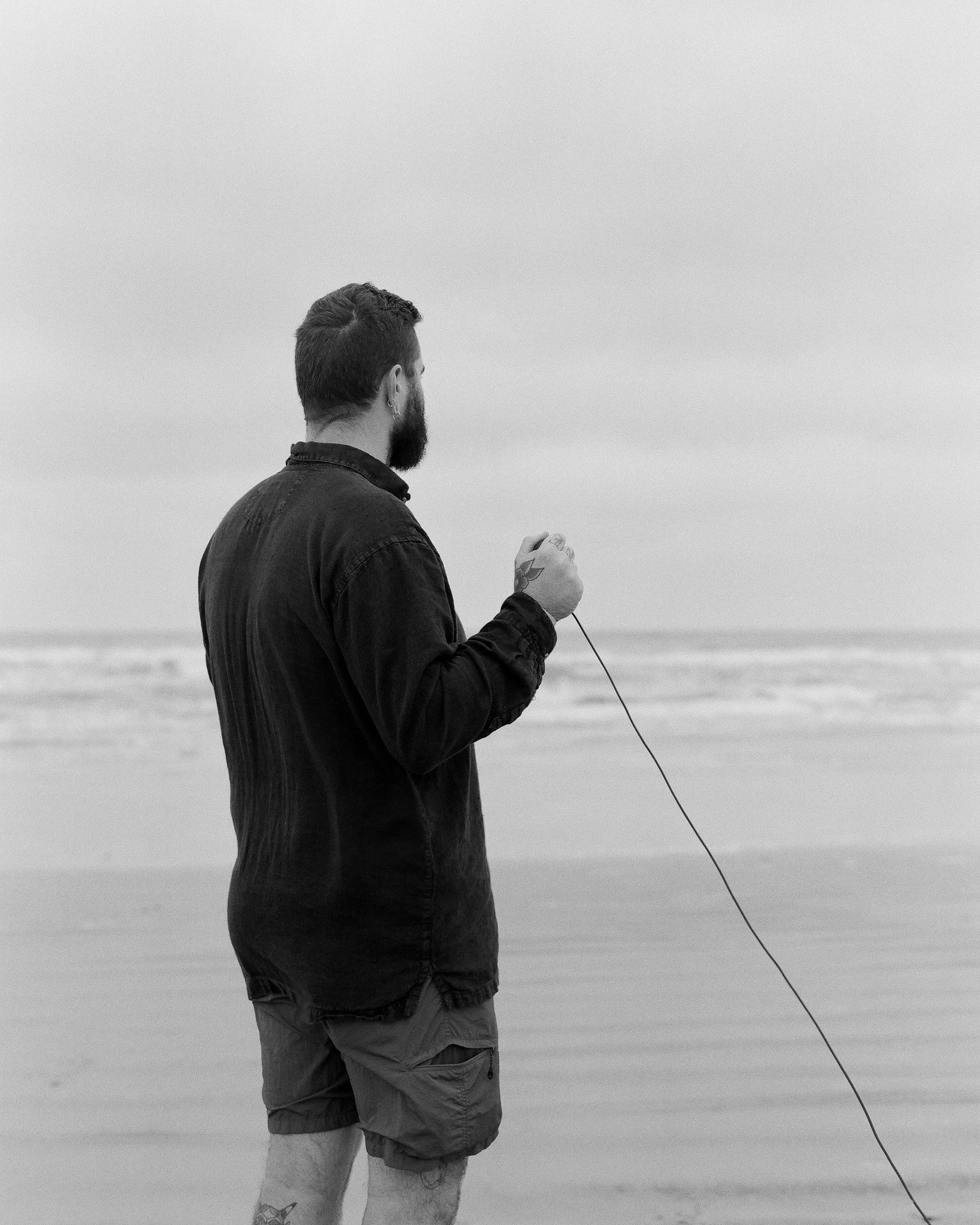 A man with a beard and tattooed legs stands on a beach, holding a fishing line while looking out at the ocean.