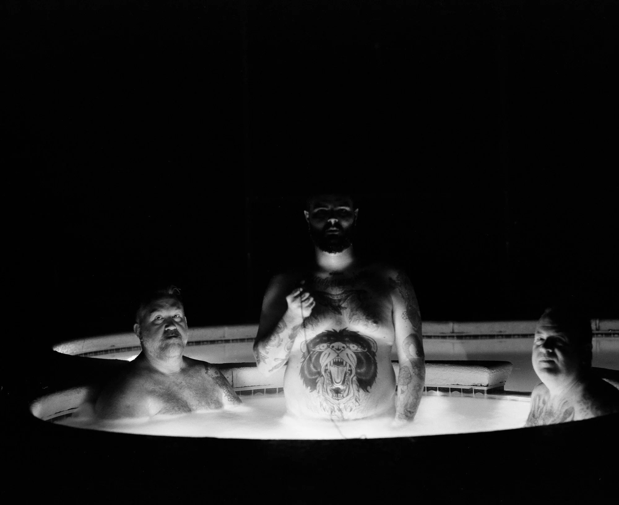 Three tattooed men are in a hot tub at night, illuminated by a single bright light.