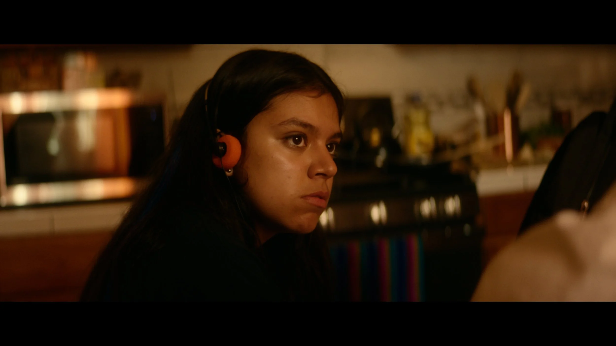 A young woman with dark hair wearing orange headphones, looking intently at a computer screen in a kitchen.