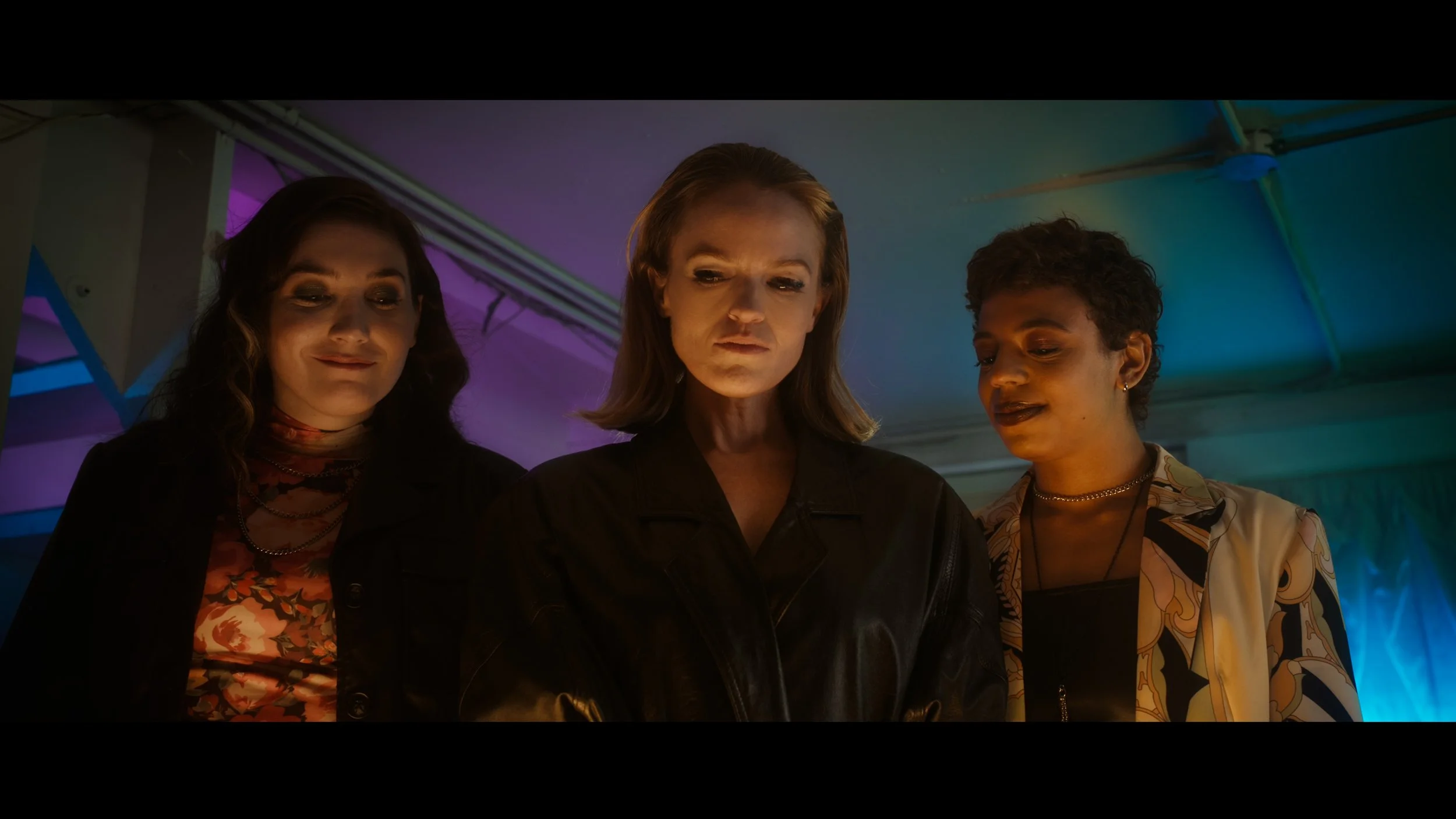 Three women standing close together indoors, looking down at something with serious and contemplative expressions, illuminated by colored lights.